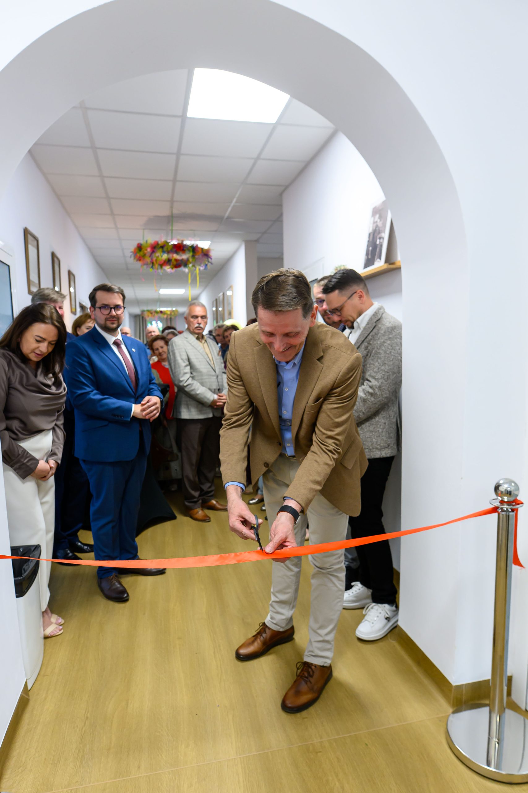 Mężczyzna w brązowej marynarce przecina czerwoną wstęgę w jasnym korytarzu. Wokół niego stoi grupa elegancko ubranych osób. Tło jest białe z drewnianą podłogą, dekoracje w tle.