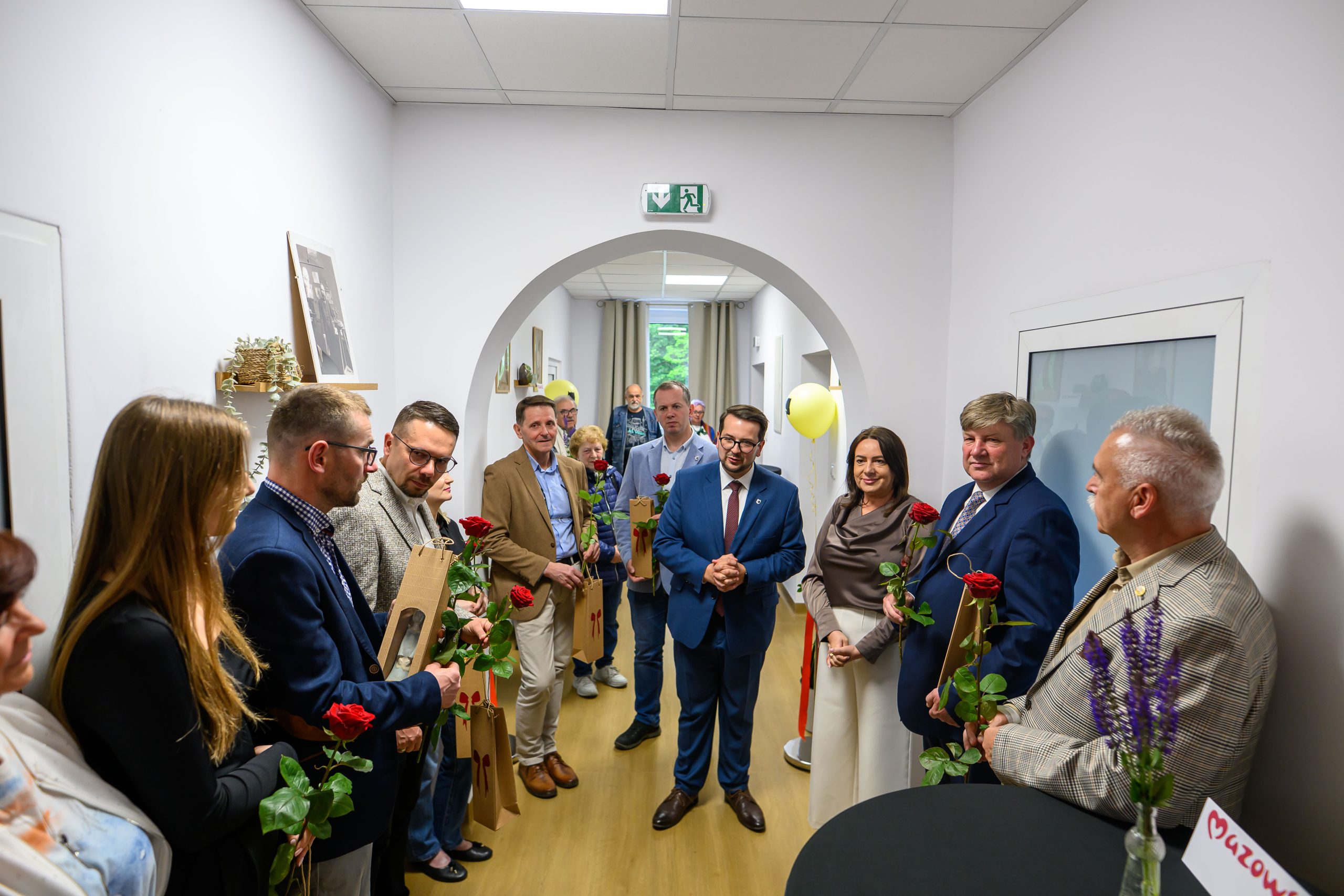 Grupa ludzi stoi w korytarzu, trzymając róże i torby. Ściany są jasne, a na stole widać lawendę. W tle znajdują się balony i wiszące zdjęcia.