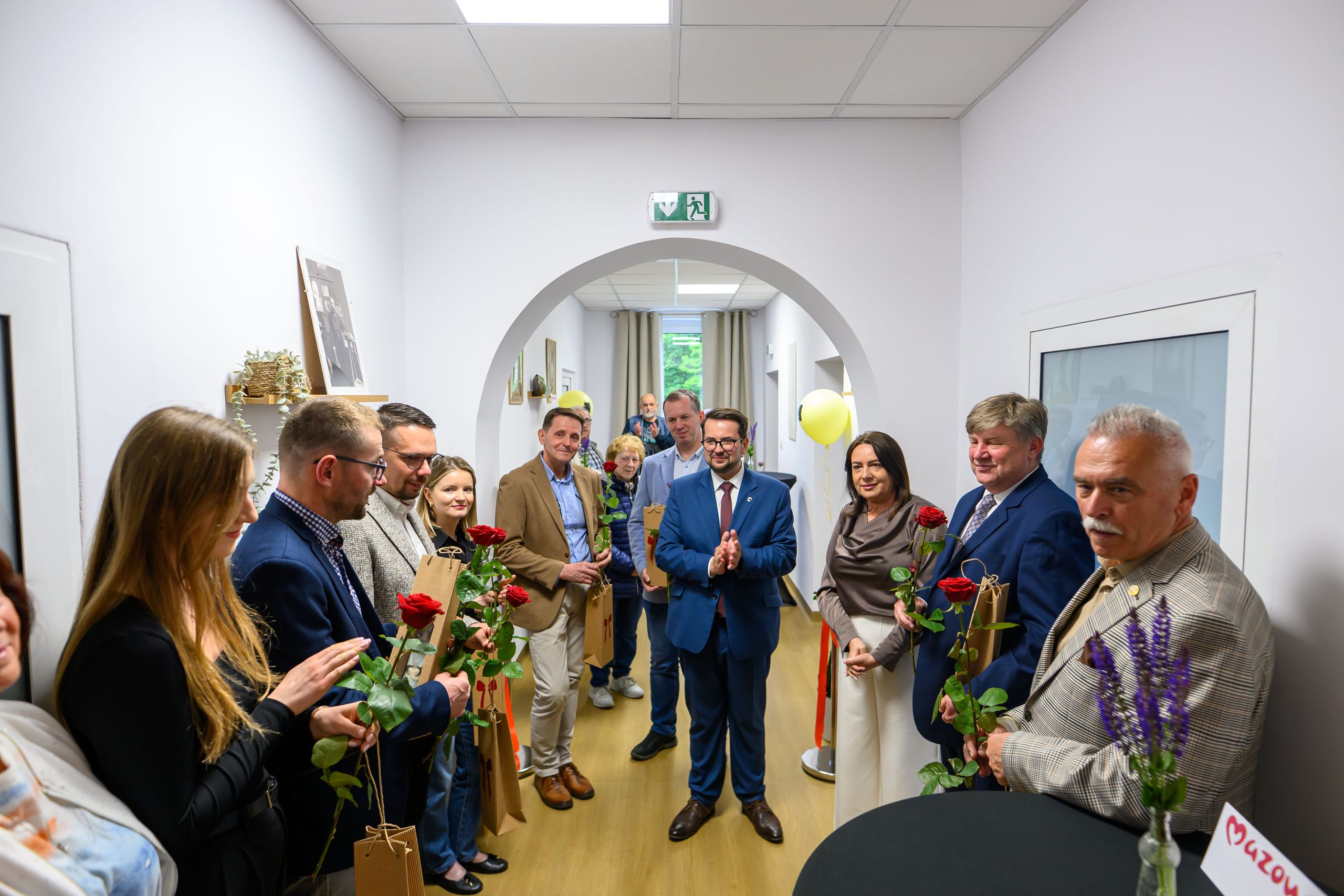 Grupa ludzi w korytarzu z drewnianą podłogą trzyma czerwone róże. Ściany są białe, a w tle widoczne są rośliny i balony. Osoby są elegancko ubrane, stoją w półkolu, patrząc w stronę kamery