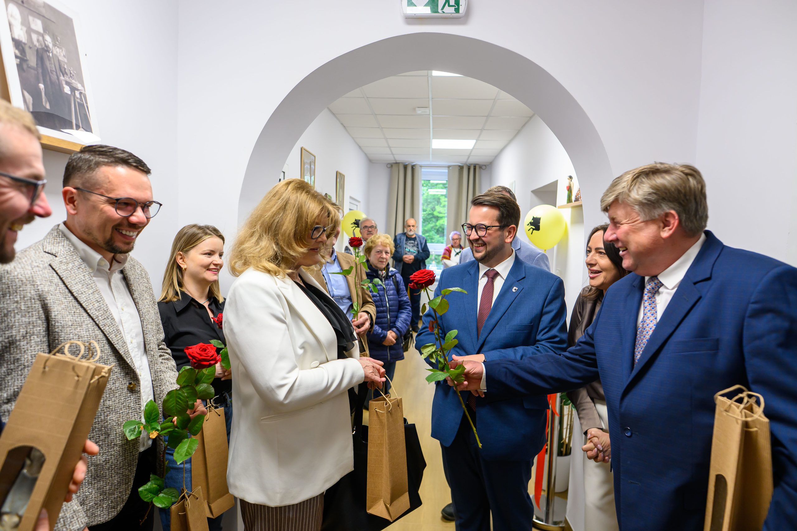 Grupa osób w eleganckich strojach w jasnym wnętrzu wymienia róże i uśmiechy. Tło z jasnymi ścianami i obrazami. Obecne są balony dekoracyjne.