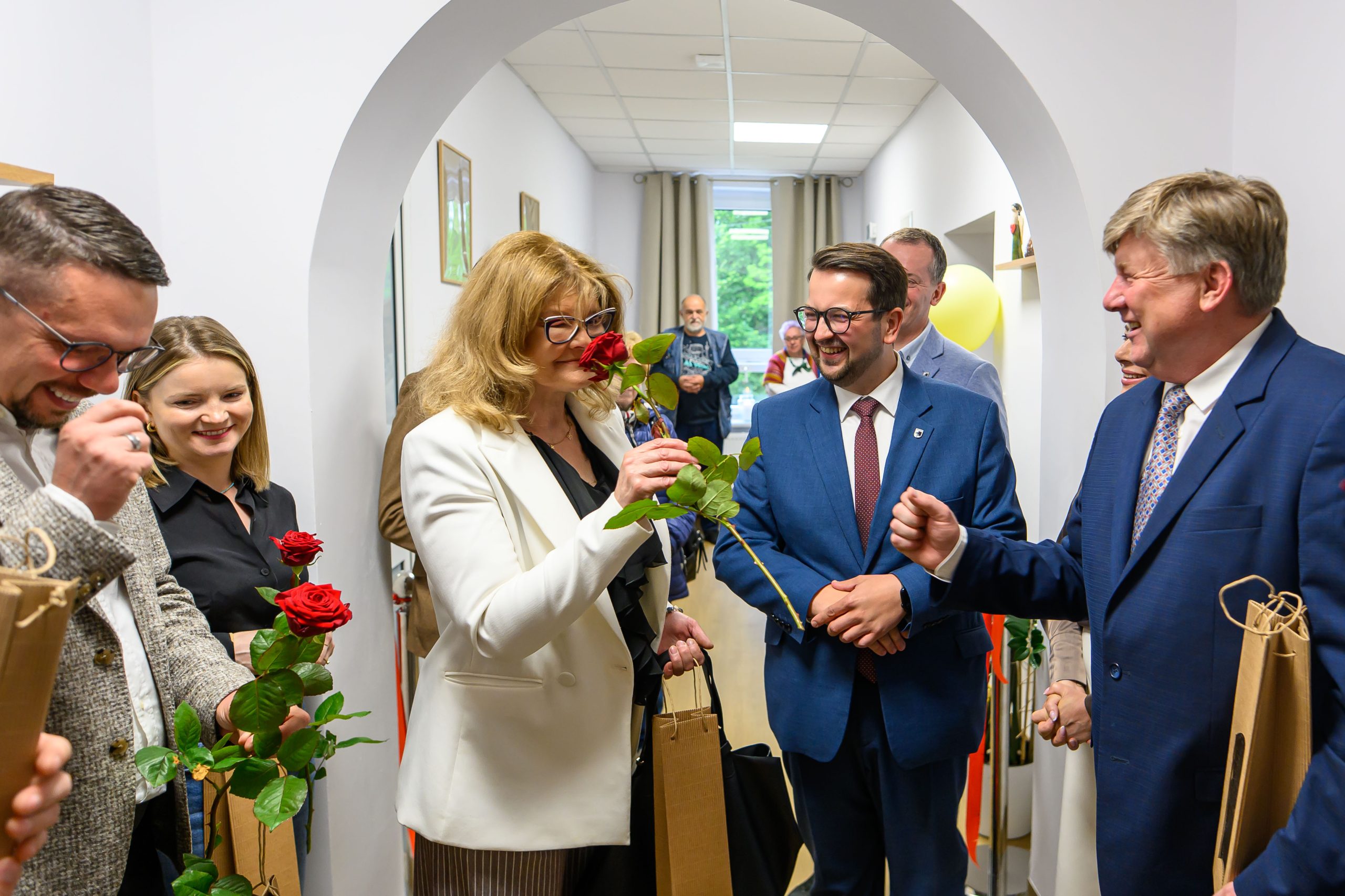 Grupa ludzi w eleganckich strojach stoi w jasnym pomieszczeniu, trzymając czerwone róże. Tło z białymi ścianami i obrazkami. Uśmiechy sugerują uroczyste wydarzenie.