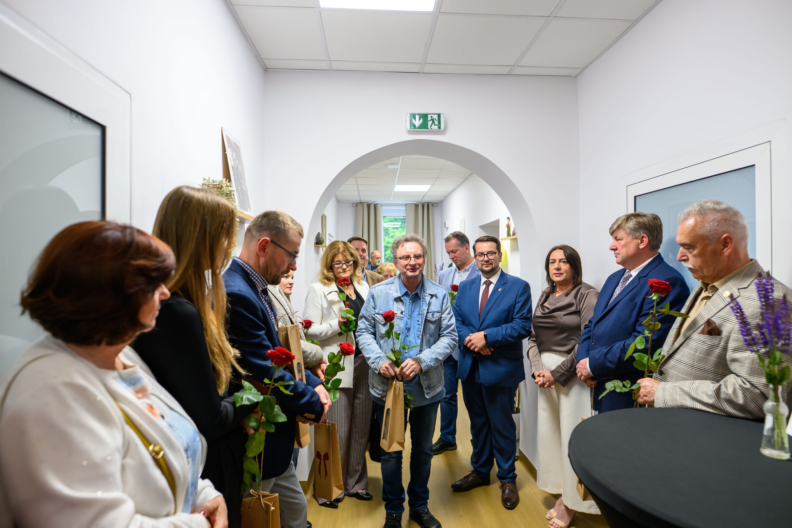 Na zdjęciu widoczna jest grupa osób stojących w korytarzu i trzymających czerwone róże. W tle jasne ściany i okno, tło jest stonowane. Na środku osoby, jedna w dżinsowej kurtce.
