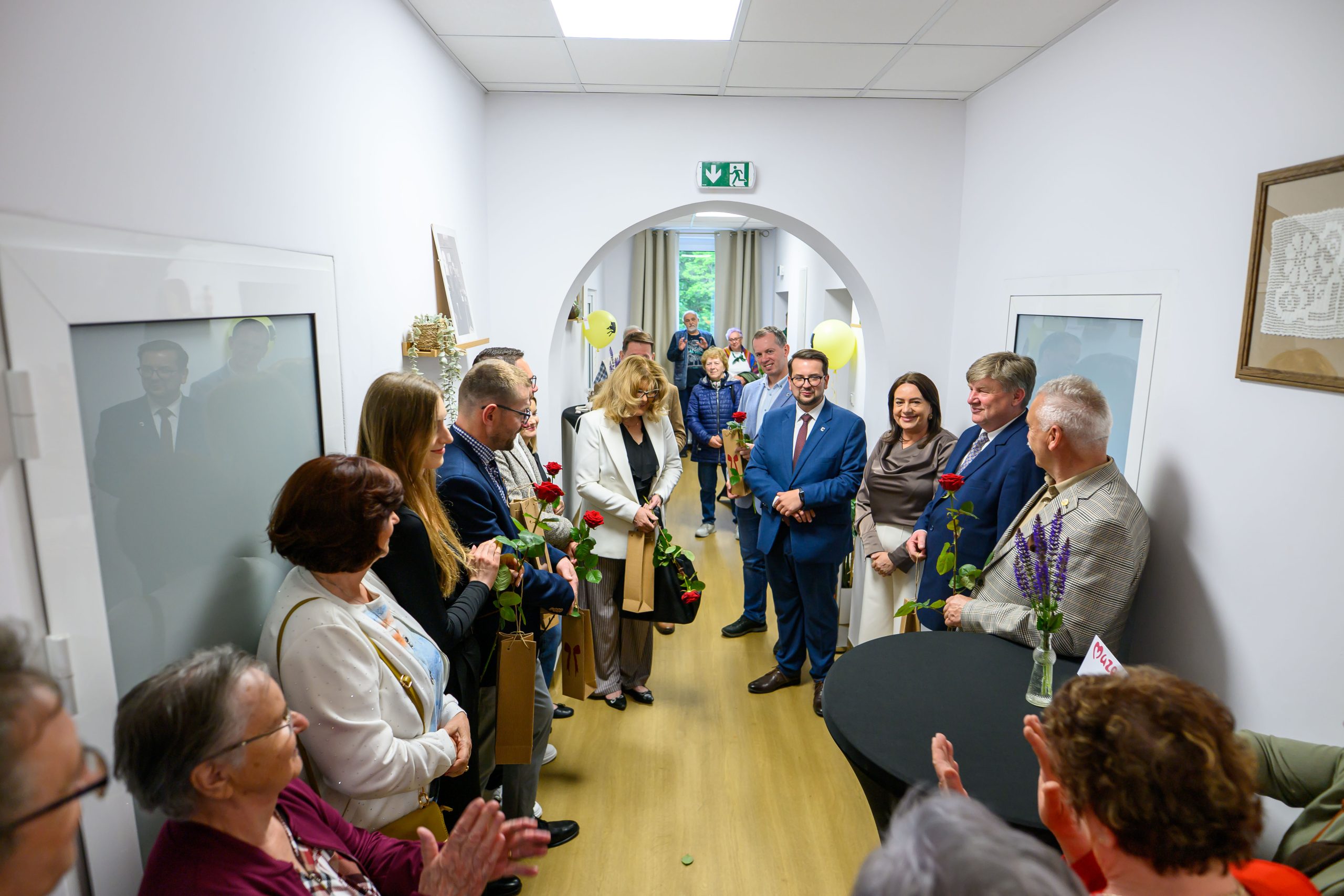 Na zdjęciu grupa osób w korytarzu, ubranych elegancko, trzyma czerwone róże. Jasne ściany i drewniana podłoga. Nastroje są radosne.
