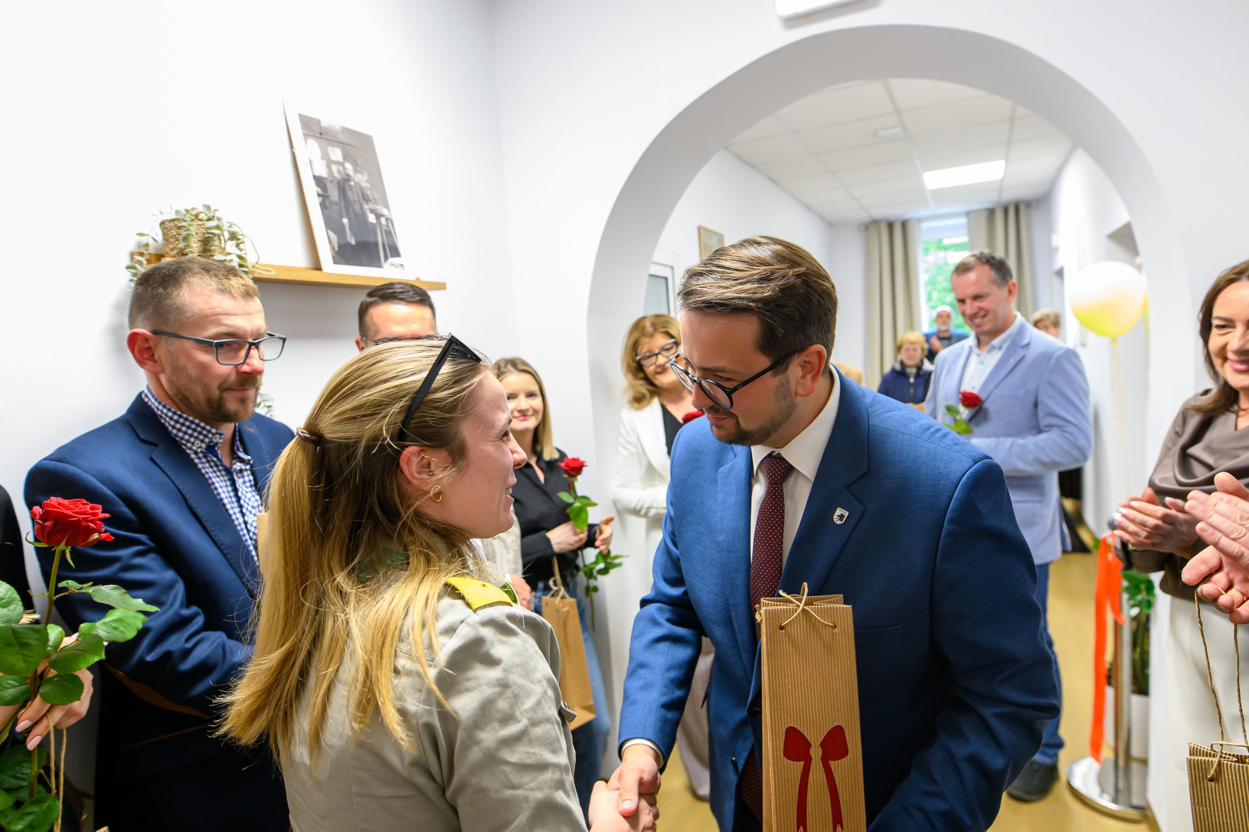 Na zdjęciu widać grupę ludzi podczas spotkania w jasnym pomieszczeniu. Osoba w niebieskiej marynarce wymienia uścisk dłoni z kobietą o blond włosach, oboje trzymają torby. W tle inni uczestnicy trzymają czerwone róże i patrzą z u