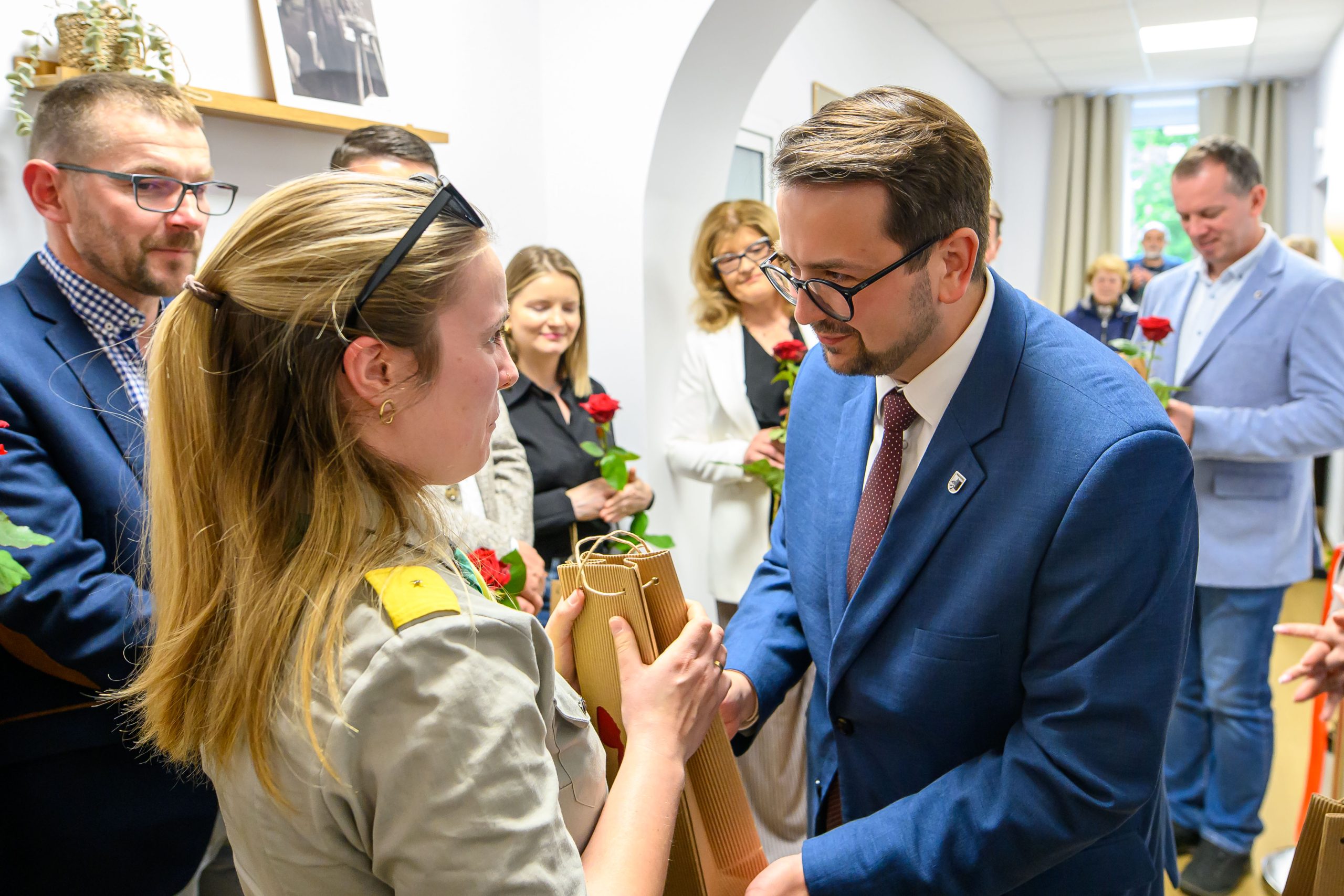 Grupa ludzi w eleganckich strojach wymienia upominki w jasnym pokoju. Kobieta trzyma różę i prezent. Mężczyzna w niebieskiej marynarce wręcza pakiet.