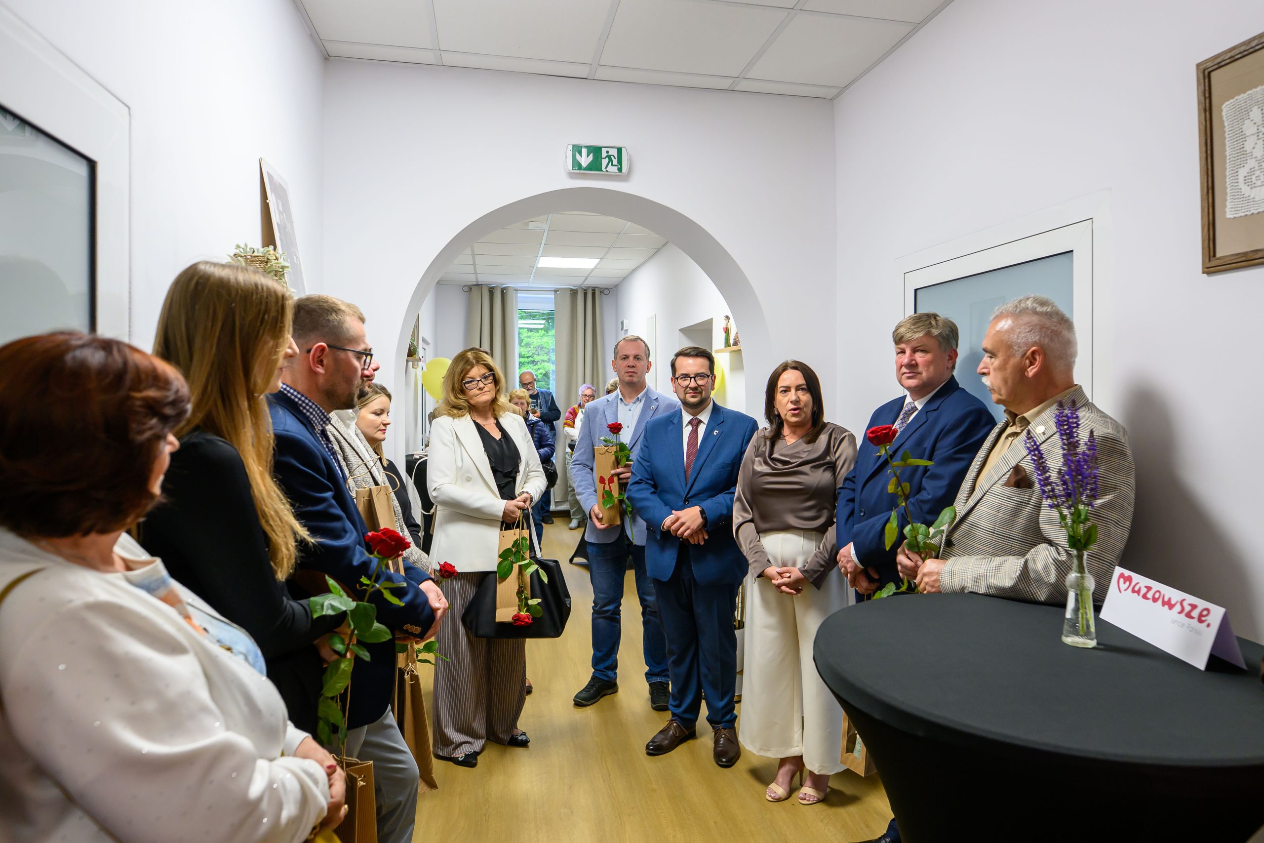 Grupa osób stoi w korytarzu, trzymając czerwone róże. Tło jest jasne, z białymi ścianami i jasną podłogą. Na stoliku obok jest tabliczka z napisem “Mazowsze”.