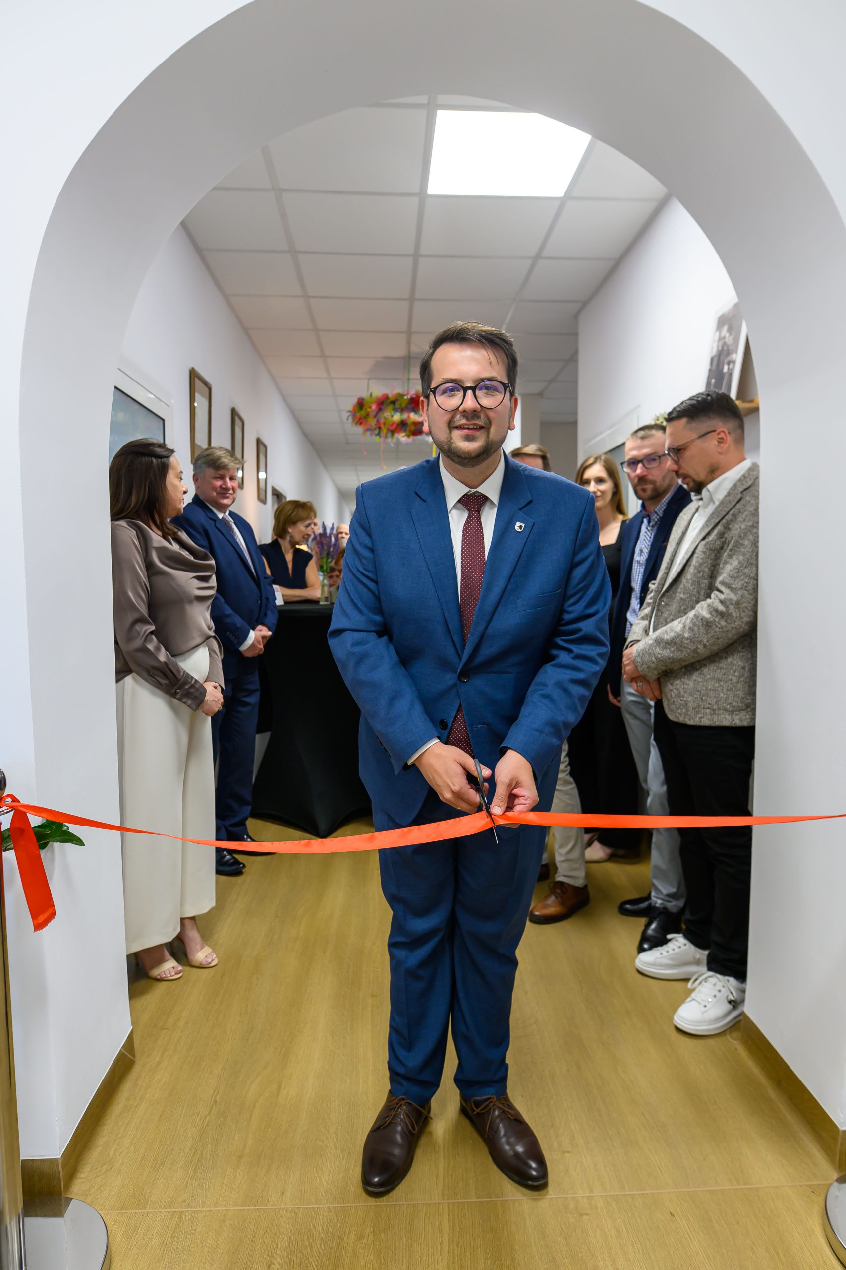Mężczyzna w niebieskim garniturze przecina czerwoną wstęgę. Stoi w jasnym korytarzu, wokół inni formalnie ubrani ludzie. W tle białe ściany i kwiatowa dekoracja.