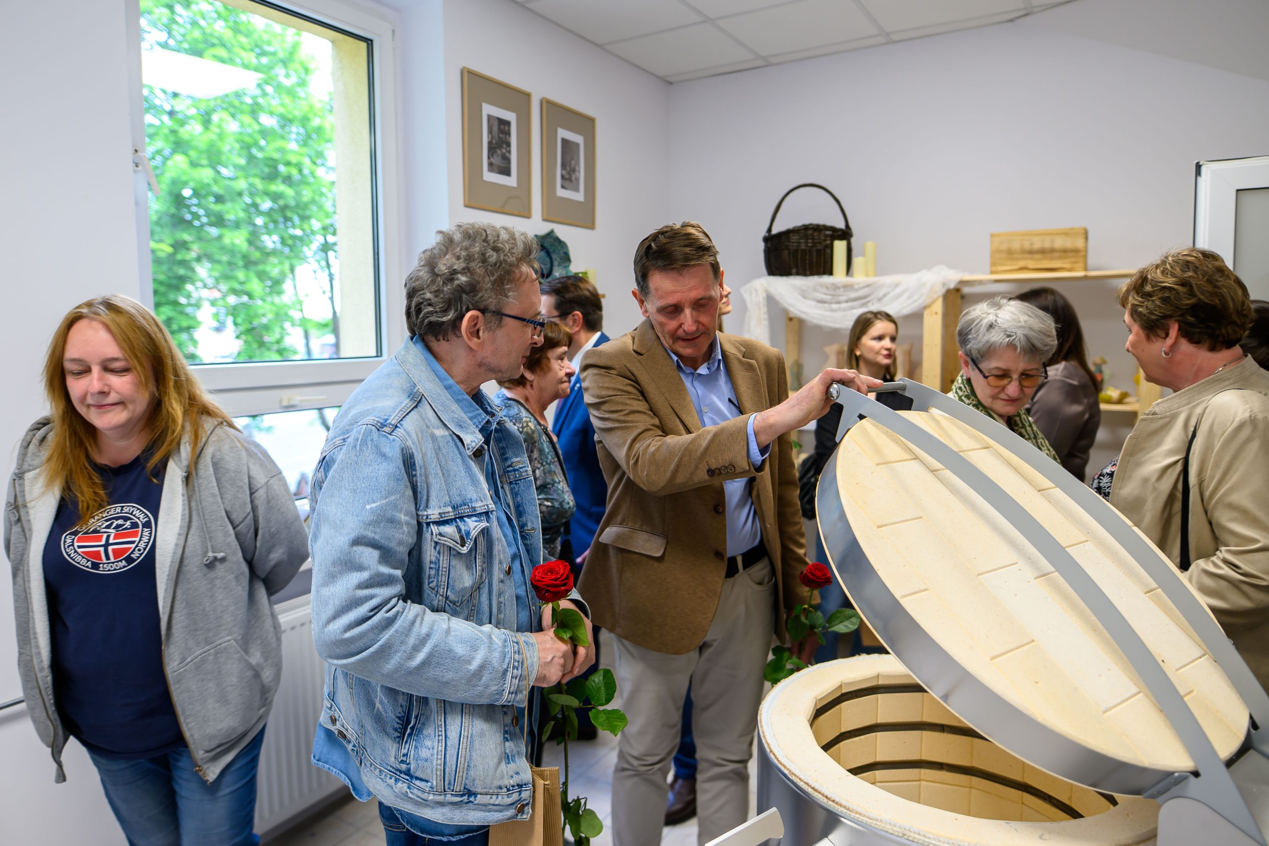 Grupa osób w jasnym pomieszczeniu ogląda otwarty piec ceramiczny. Tło stanowi ściana z oknem i obrazami. Niektórzy trzymają czerwone róże.