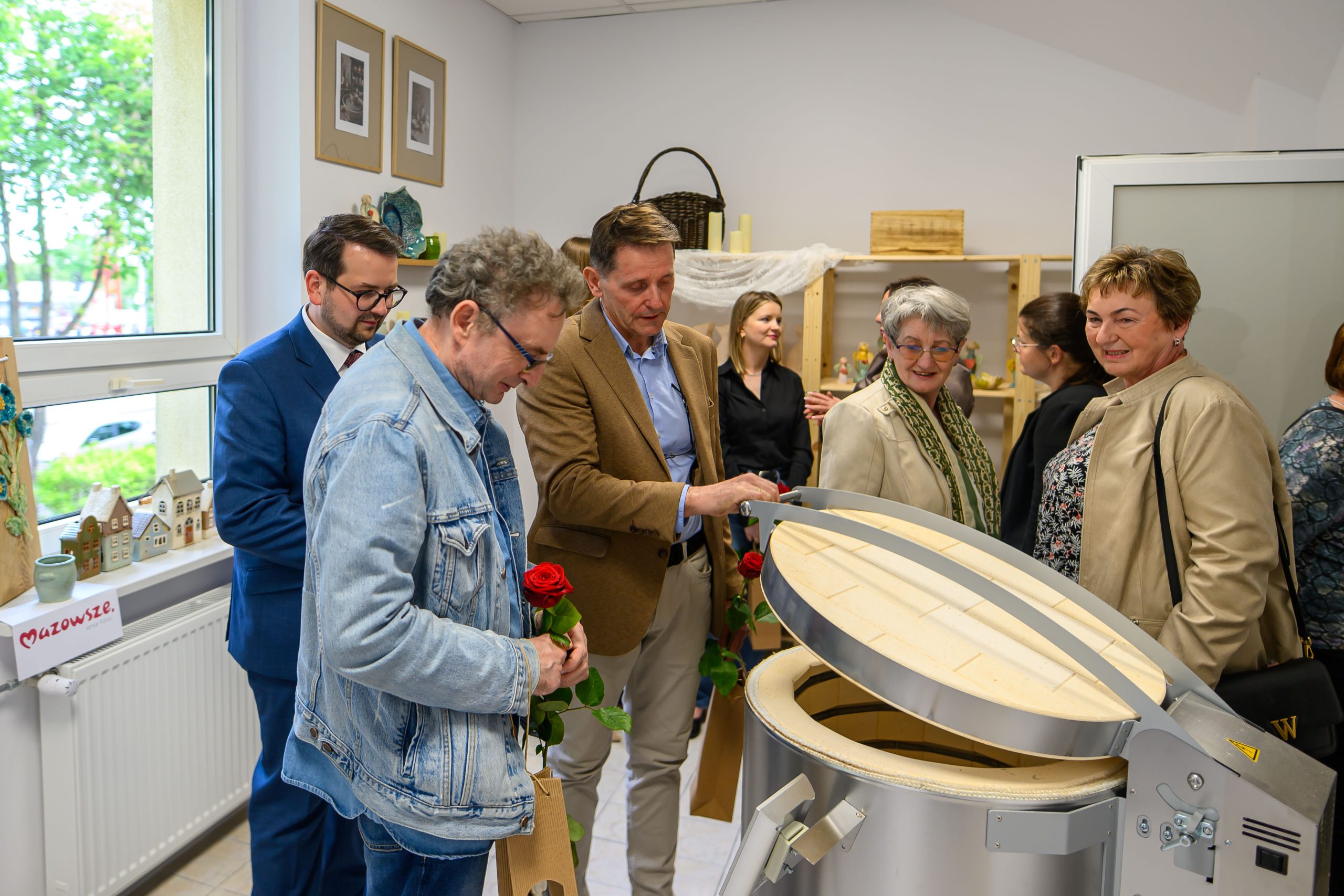 Grupa osób stoi wokół nowoczesnego pieca ceramicznego. Ubrania w stonowanych kolorach, wnętrze jasne. Przez okno widać zielone drzewa.