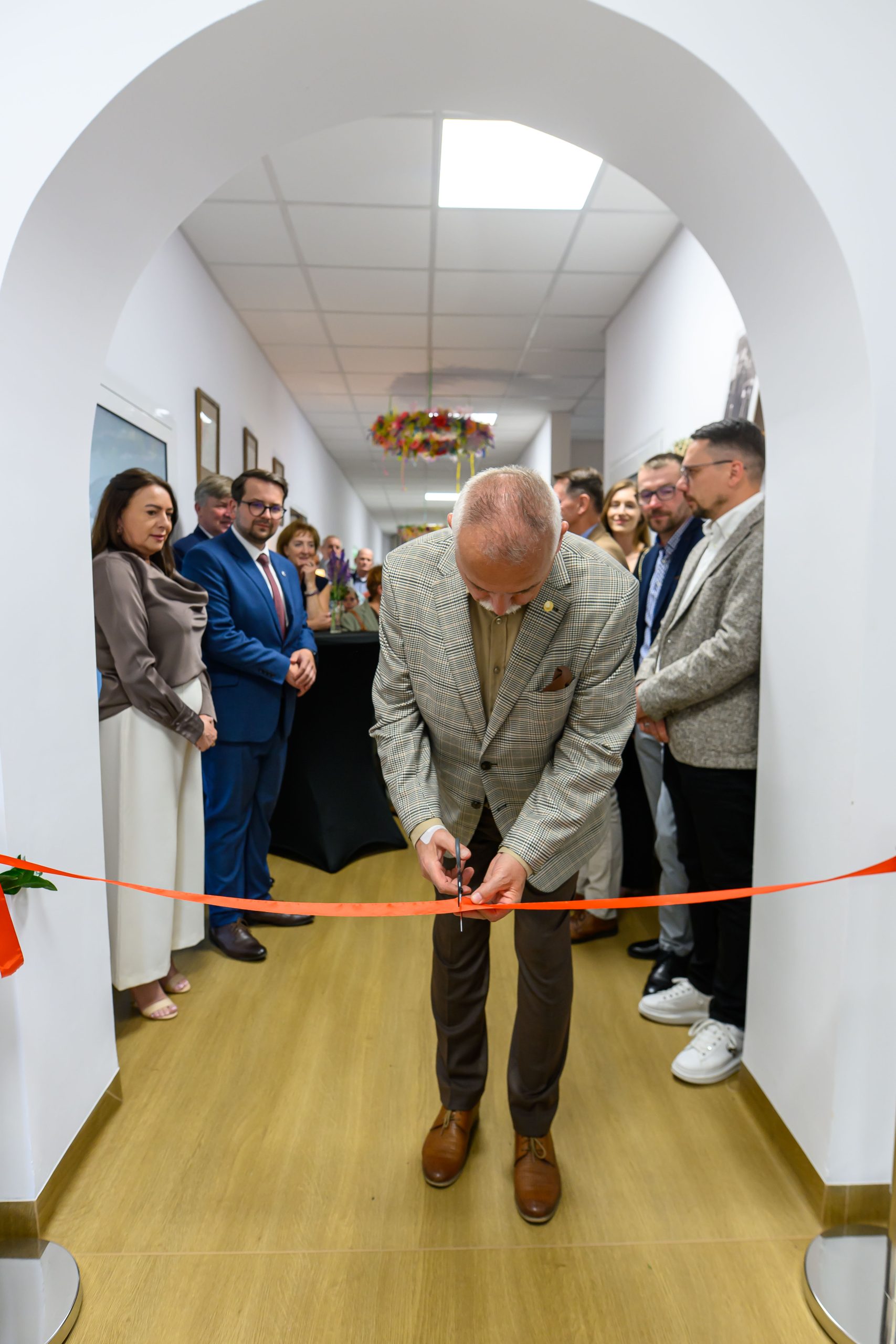 Mężczyzna w beżowej marynarce przecina czerwoną wstęgę. W tle grupa osób w formalnych strojach. Jasne, nowoczesne wnętrze.
