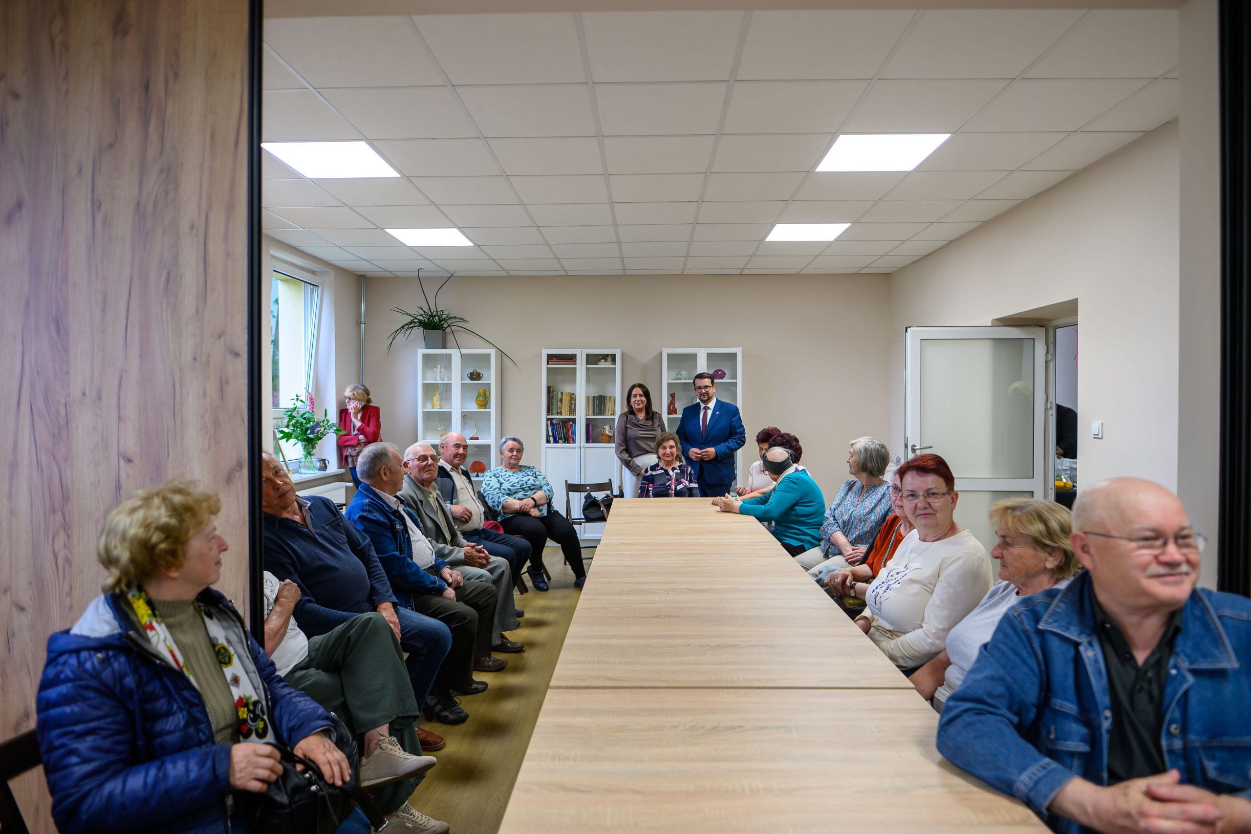 Długi stół w sali z jasnym oświetleniem. Grupa osób siedzi i stoi wokół, z roślinami i regałami w tle. Dominują beże i błękity.