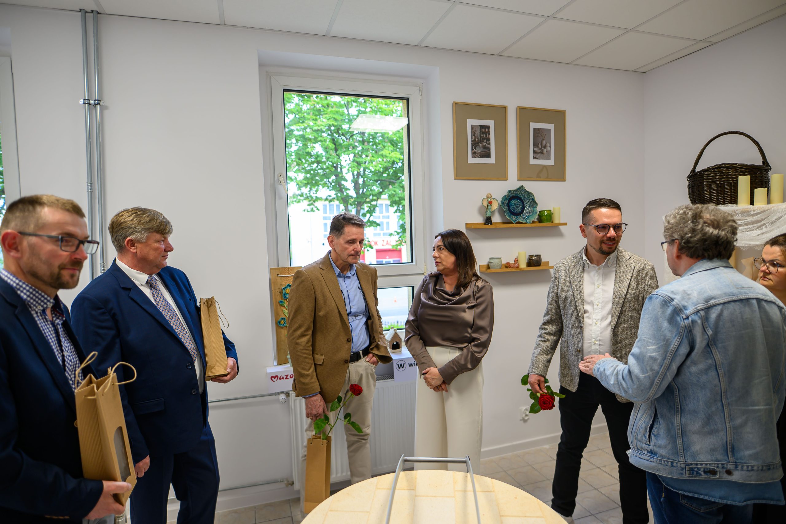 Na zdjęciu widać grupę osób w pomieszczeniu o białych ścianach. Niektórzy z nich trzymają czerwone róże i torby. W tle znajdują się półki z ozdobami.