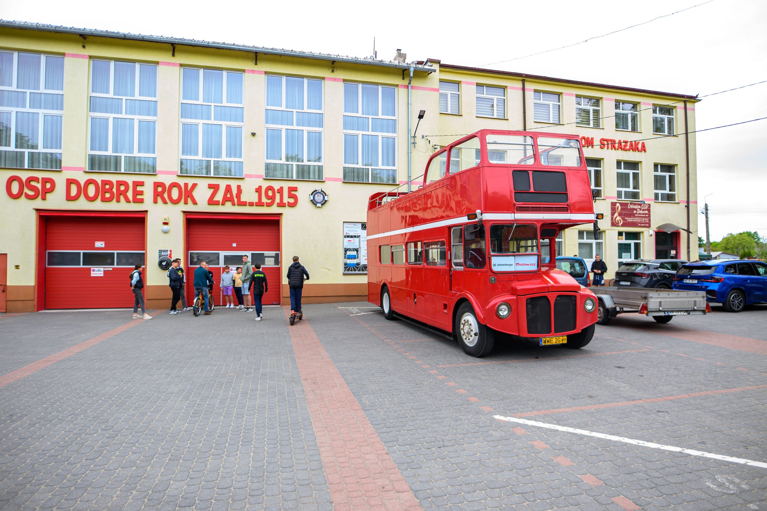 Czerwony piętrowy autobus stoi przed budynkiem straży pożarnej z napisem OSP Dobre Rok Zał. 1915. Grupa ludzi obok pojazdu. Pogodne niebo i brukowane podłoże.