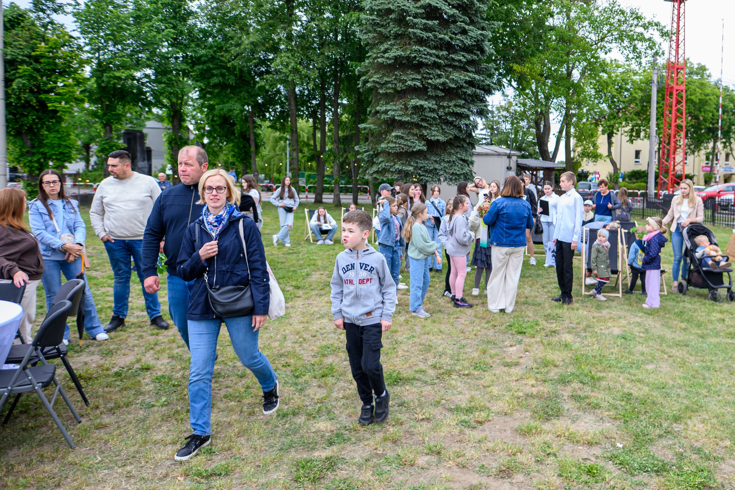 Grupa ludzi na zielonej trawie w parku. Drzewa w tle, jasne ubrania uczestników. Rodzinna atmosfera eventu plenerowego.