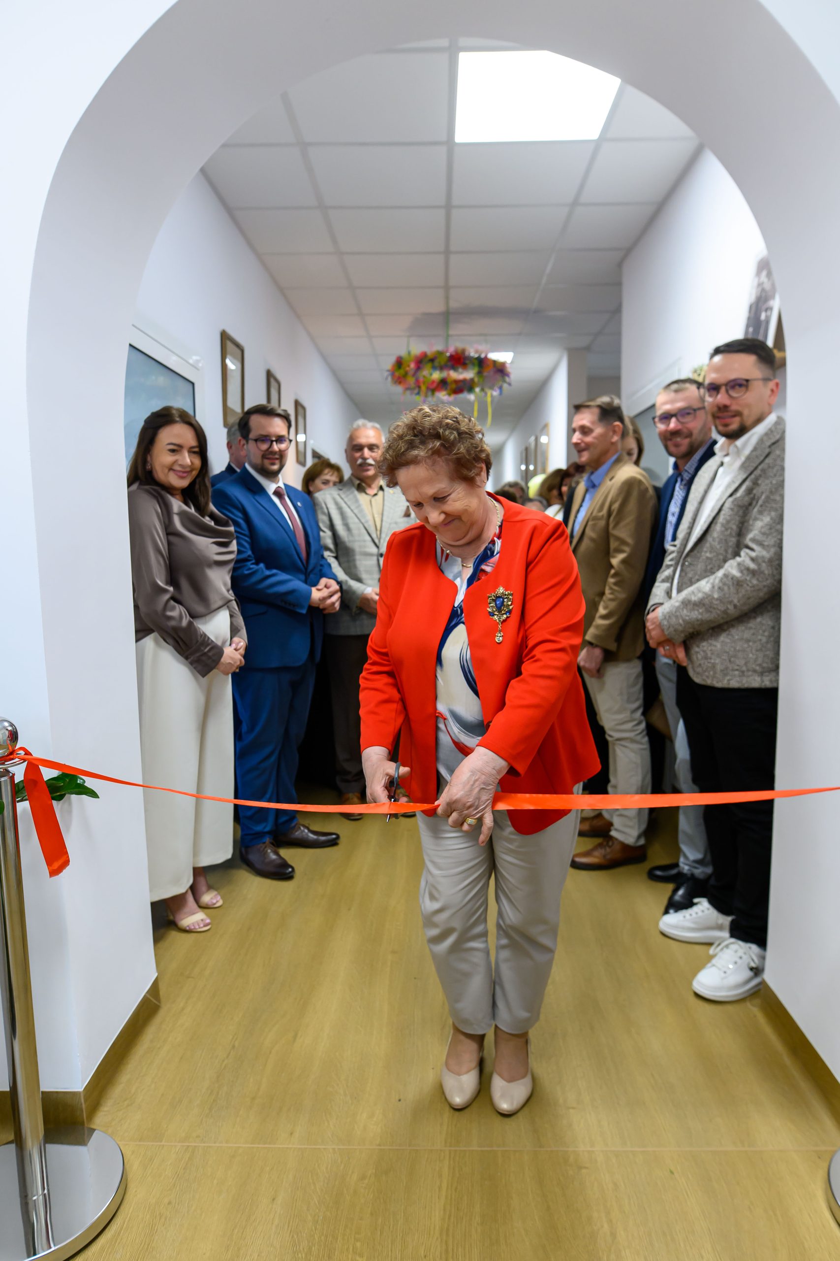Starsza kobieta w czerwonej marynarce przecina czerwoną wstęgę w jasnym korytarzu, otoczona przez grupę ludzi.