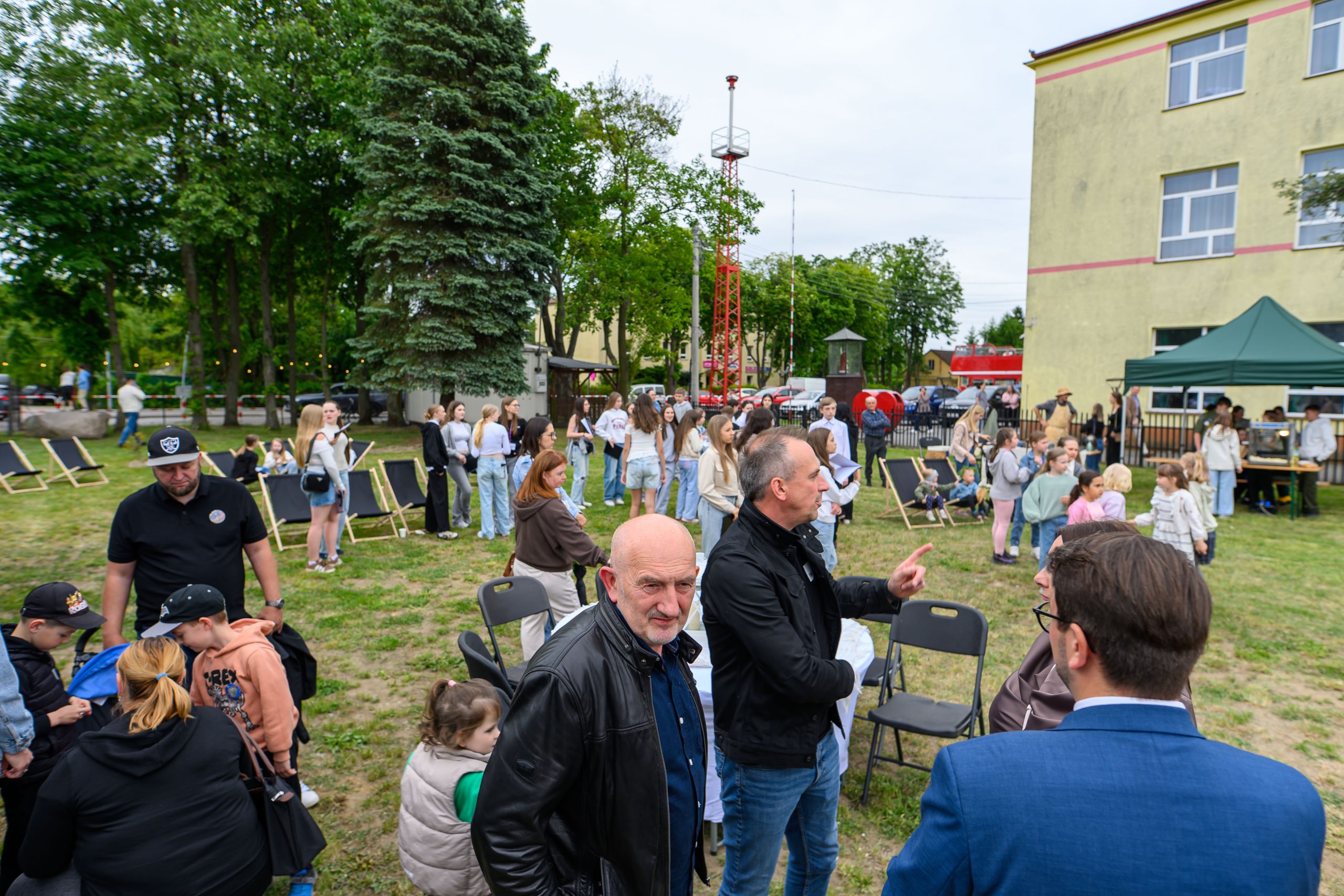 Grupa ludzi na trawniku przed budynkiem. Zielone drzewo z lewej, namiot z prawej. Dzieci i dorośli rozmawiają.