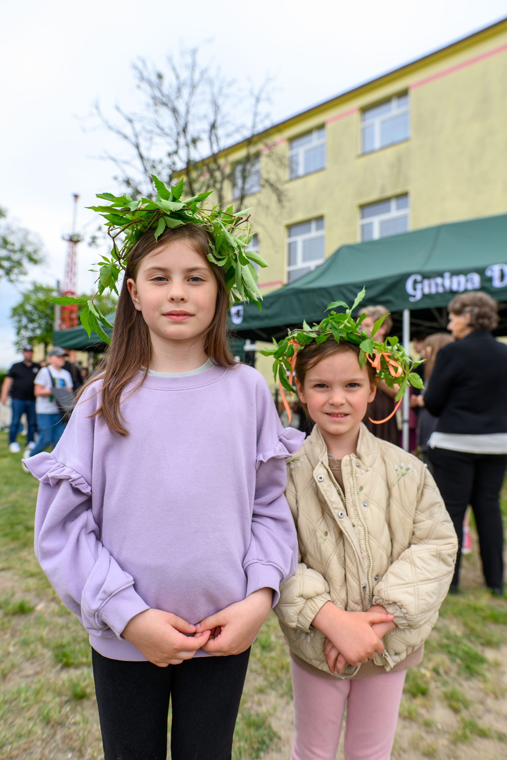 Dwie dziewczynki w wiankach z liści stoją na tle zielonego namiotu i żółtego budynku. Jedna ma fioletowy sweter, druga beżową kurtkę. W tle grupka osób.