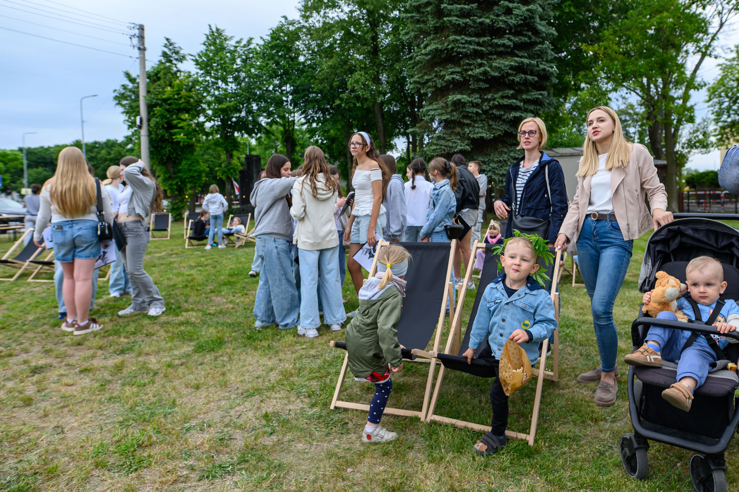 Grupa ludzi na zielonej trawie; dzieci, młodzież i dorośli w casualowych ubraniach, w tle drzewa i niebo.