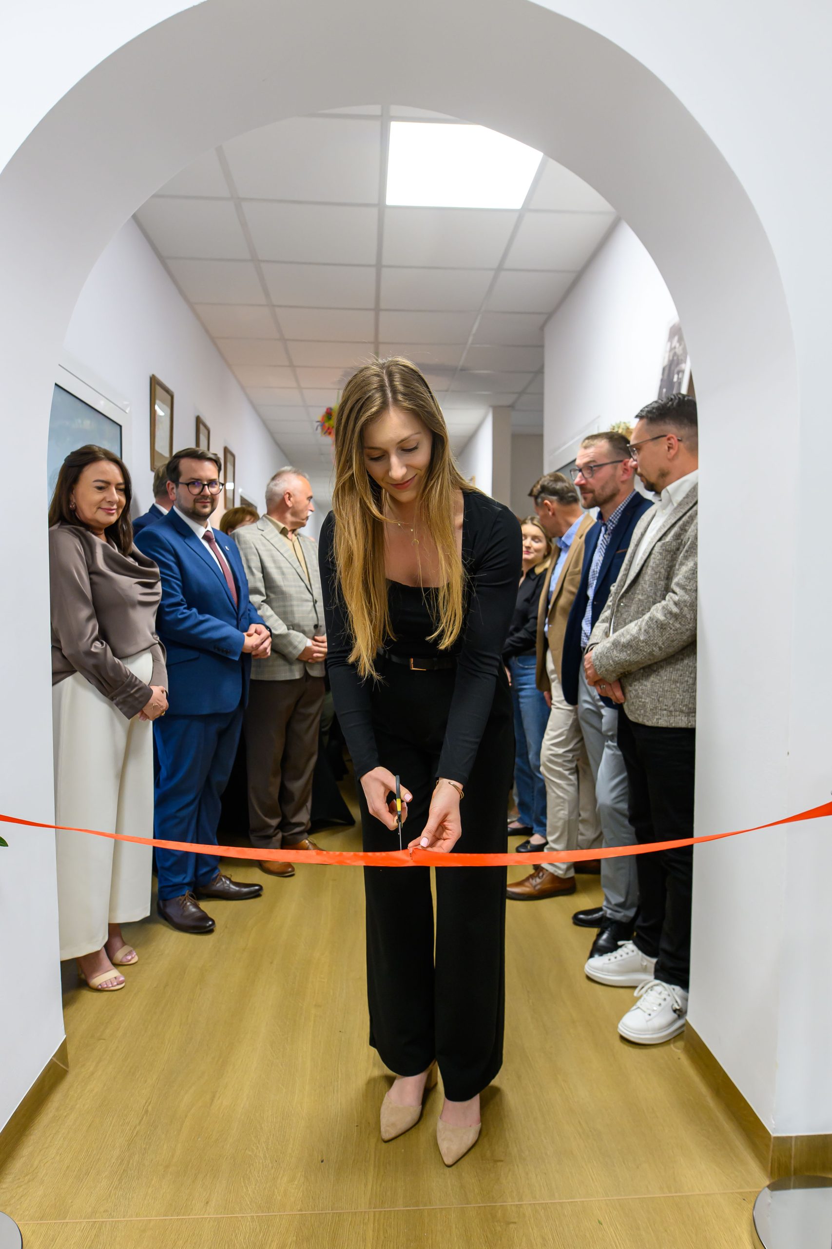 Kobieta w czarnym stroju przecina czerwoną wstęgę w białym korytarzu. W tle grupa elegancko ubranych osób. Wszystko odbywa się w jasnym, przestronnym pomieszczeniu.
