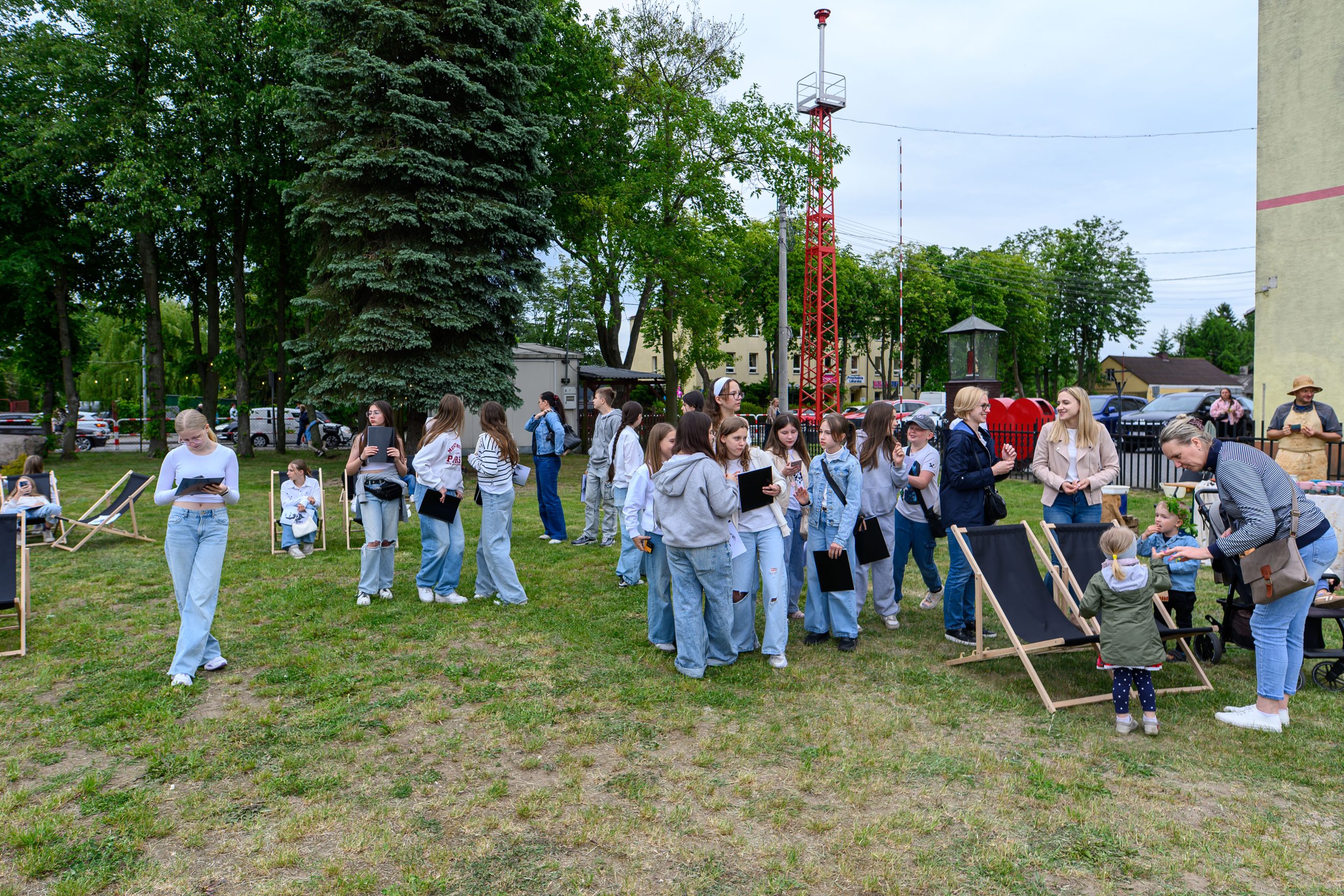 Grupa ludzi na trawie w parku, głównie młodzież w jasnych ubraniach. W tle drzewa i czerwona wieża. Na pierwszym planie leżaki i kobieta z dzieckiem.