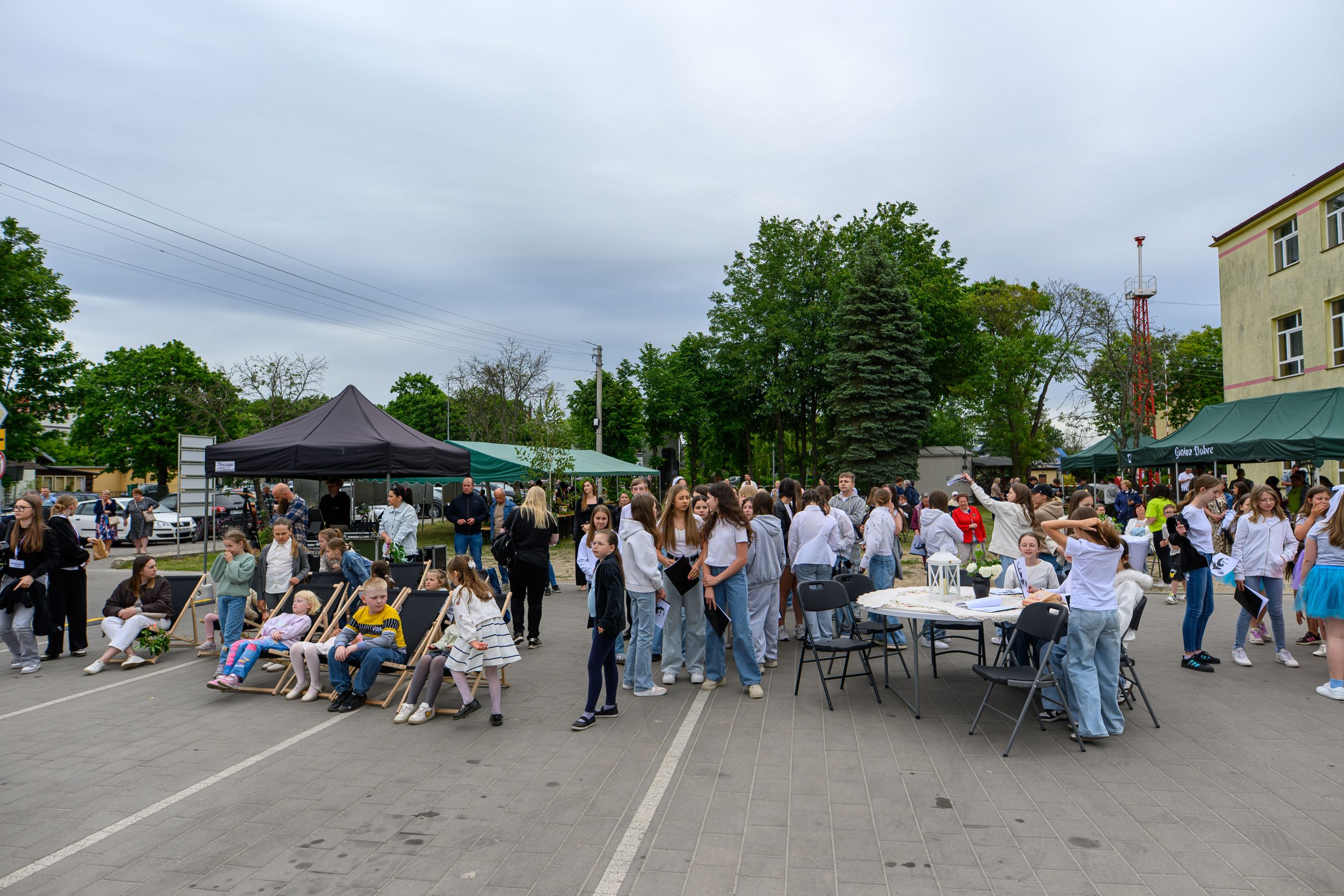 Na zdjęciu pokazane jest wydarzenie plenerowe z dużą grupą ludzi na placu. W tle są drzewa, namioty i budynek. Przeważają kolory szary, zielony i biały.