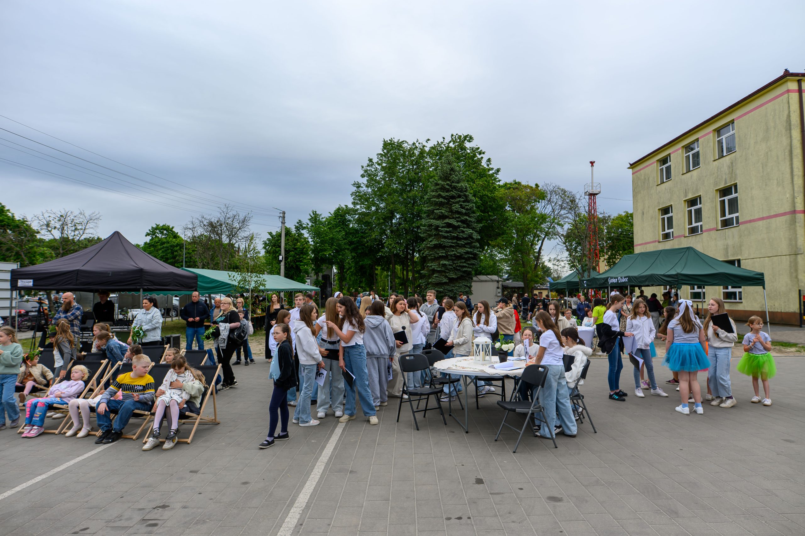 Na zdjęciu widać tłum ludzi podczas wydarzenia plenerowego. Przedstawione są kolorowe namioty, dzieci na leżakach i zielone drzewa w tle. Po prawej stronie znajduje się budynek o żółtych ścianach.