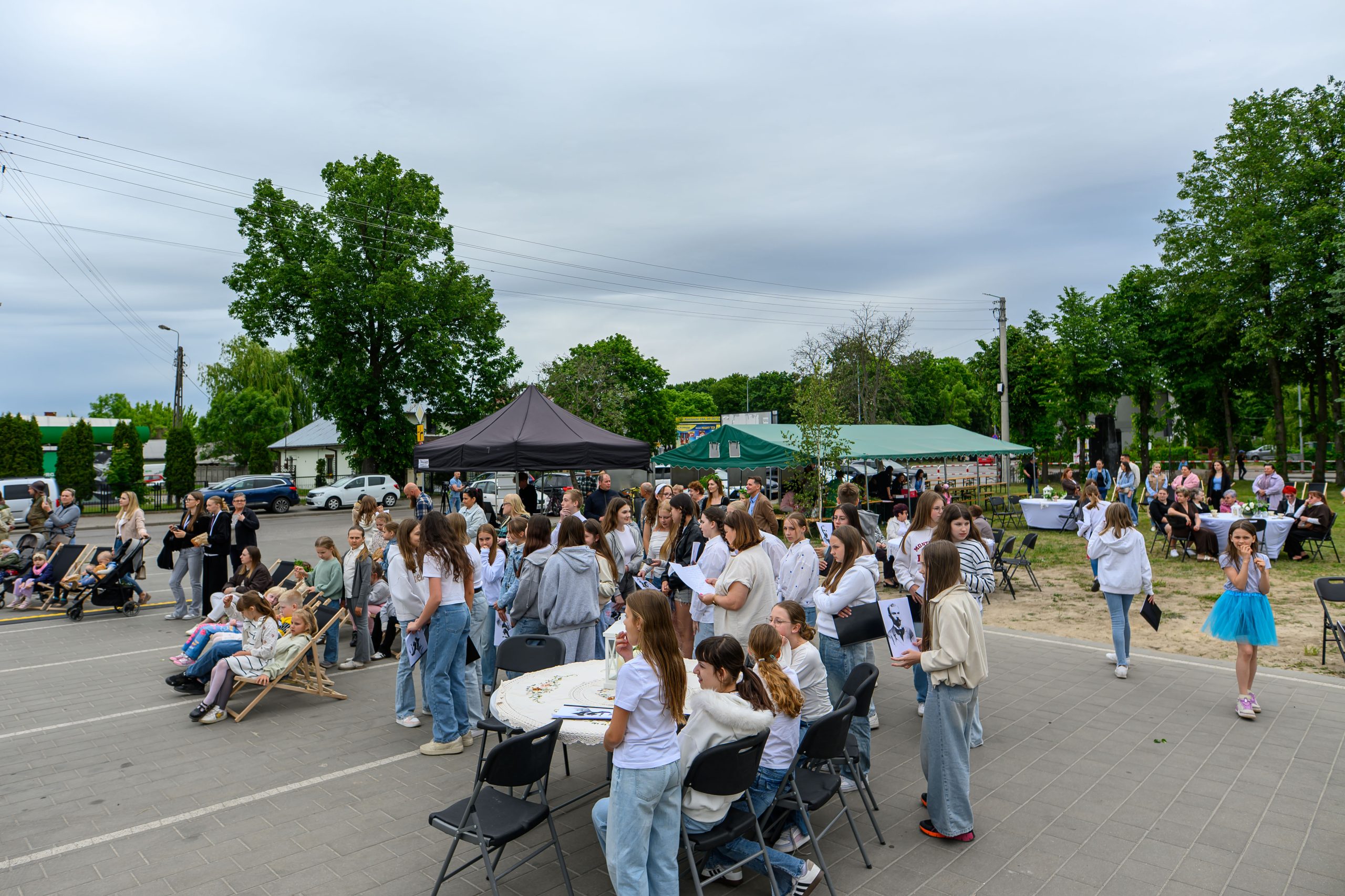 Na zdjęciu widać grupę ludzi biorących udział w plenerowym wydarzeniu. Tło stanowią zielone drzewa i namioty. Uczestnicy są w swobodnych ubraniach, atmosfera jest radosna.