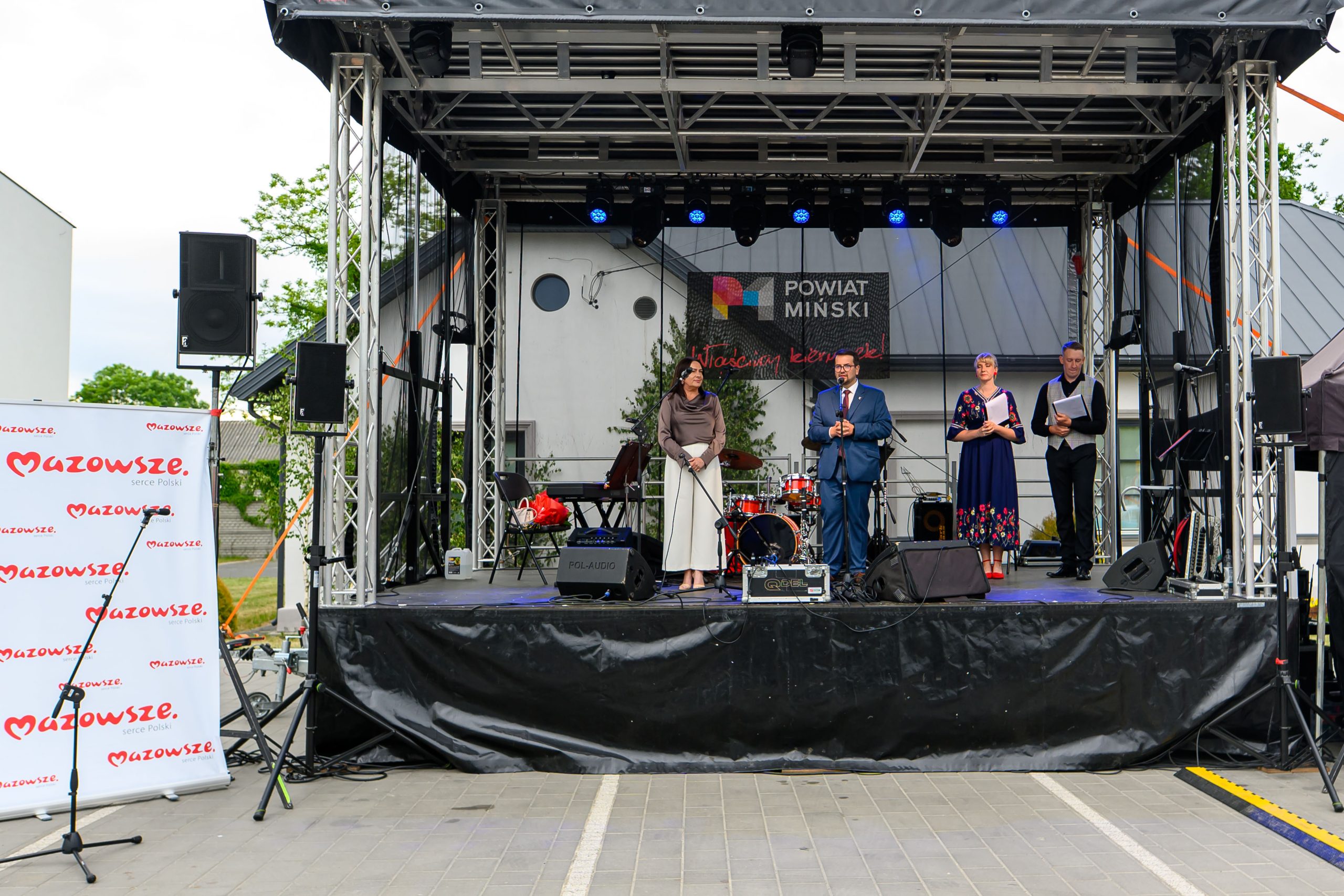 Scena plenerowa z ludźmi przemawiającymi do mikrofonów. Tło z banerem Powiat Miński. Po lewej biały roll-up Mazowsze.