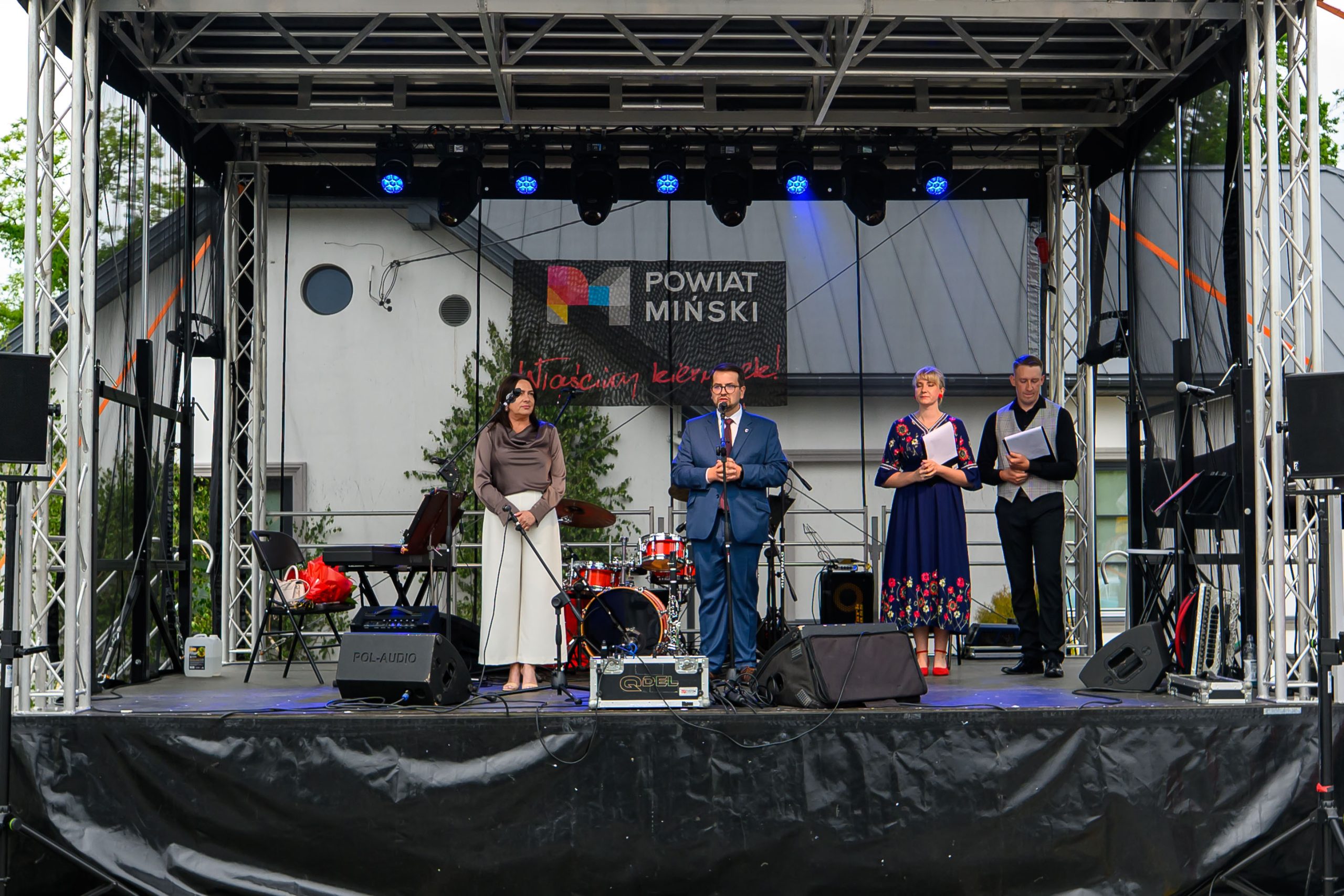 Grupa osób stoi na scenie podczas wydarzenia powiatowego. W tle widoczny jest baner z napisem Powiat Miński. Na scenie znajdują się instrumenty muzyczne i sprzęt audio.