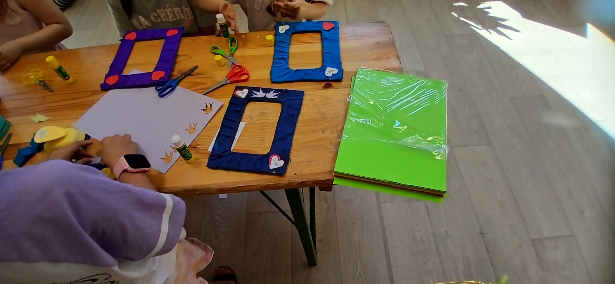 Dzieci przy stole tworzą dekoracje z papieru. Na blacie kolorowe ramki i nożyczki. Obok zielone arkusze papieru.