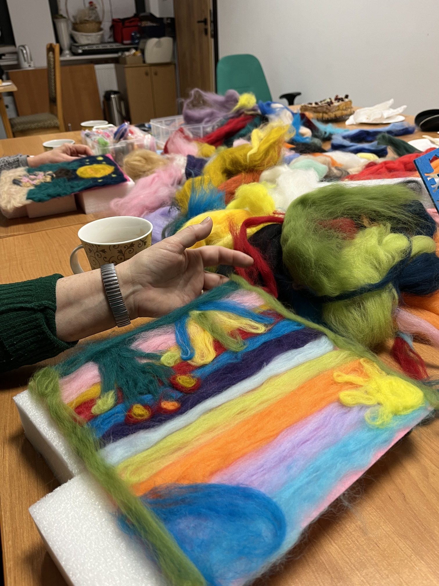 Na zdjęciu widać kolorowe wełniane włókna rozłożone na stole. Osoba trzyma jasną tkaninę z zielonym wzorem. W tle są różne przybory i filiżanki.