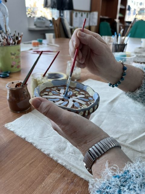 Osoba maluje małą ceramikę w oryginalny wzór, używając pędzla. Kolory jasnobrązowy, niebieski i biały dominują na tle drewnianego stołu. W tle widoczne są przybory malarskie oraz półki.