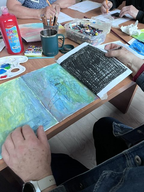 Osoba rysuje w szkicowniku przy stole pełnym kredek, farb i innych materiałów plastycznych. Kolory to głównie zielenie, czernie i błękity. Tło stanowi jasnobrązowy blat stołu z białą podłogą.