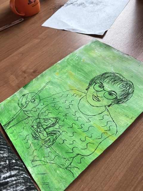 Obrazek przedstawia portret kobiety z włosami do ramion na zielonym tle. Kobieta nosi okulary i trzyma długopis. Drewniane biurko w tle.