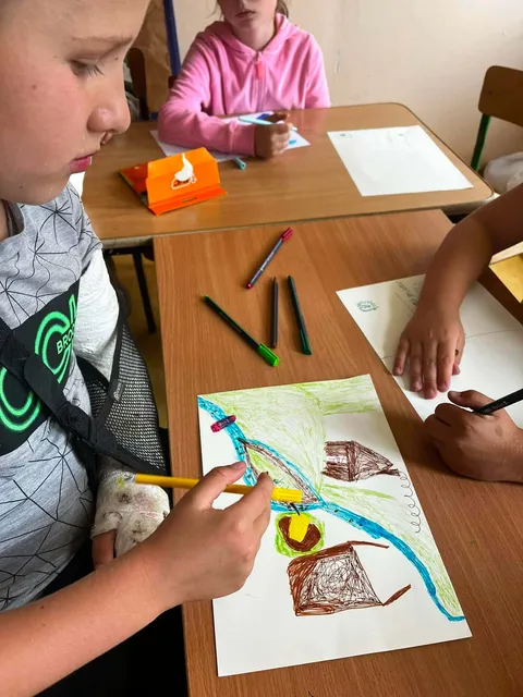 Na zdjęciu dzieci rysują na kartkach przy drewnianych stołach. Dominują żywe kolory - żółty, zielony i niebieski. W tle widać pudełko chusteczek i różowe ubranie.