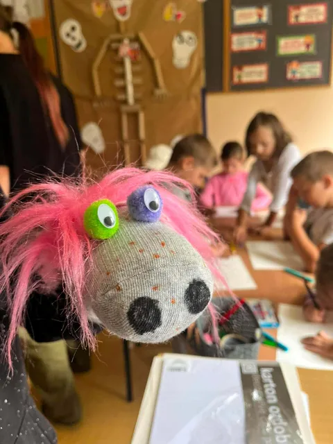 Na pierwszym planie różowa pacynka z dużymi oczami. W tle dzieci rysują przy stole, otoczone pracami plastycznymi. Kolory są jasne i żywe.