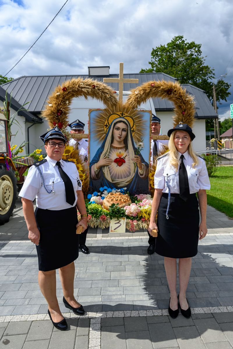 Na zdjęciu widoczne są dwie osoby w mundurach strażackich obok obrazu Matki Boskiej. Tło stanowi budynek i niebo z chmurami. Dekoracje z kwiatów i zboża dodają kolorów.