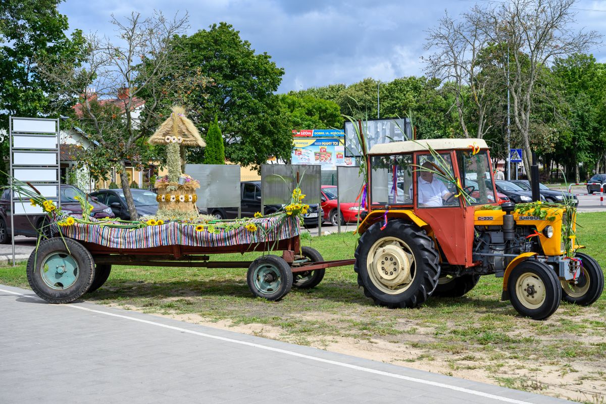 Traktor z czerwoną przyczepą ozdobioną kwiatami stoi na tle zielonych drzew i nieba. Przyczepa przystrojona kolorowymi wstążkami i sianem. Tło stanowią zaparkowane auta i tablice reklamowe.
