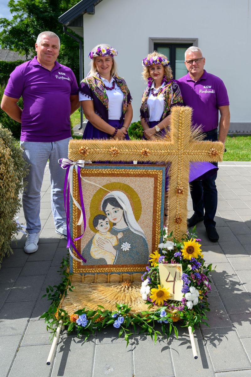 Grupa czterech osób w fioletowych koszulkach stoi za ozdobnym wieńcem z wizerunkiem Madonny z Dzieciątkiem, otoczonym kwiatami. W tle biały budynek i zieleń. Ozdoba składa się z kłosów zboża i kolorowych kwiatów.