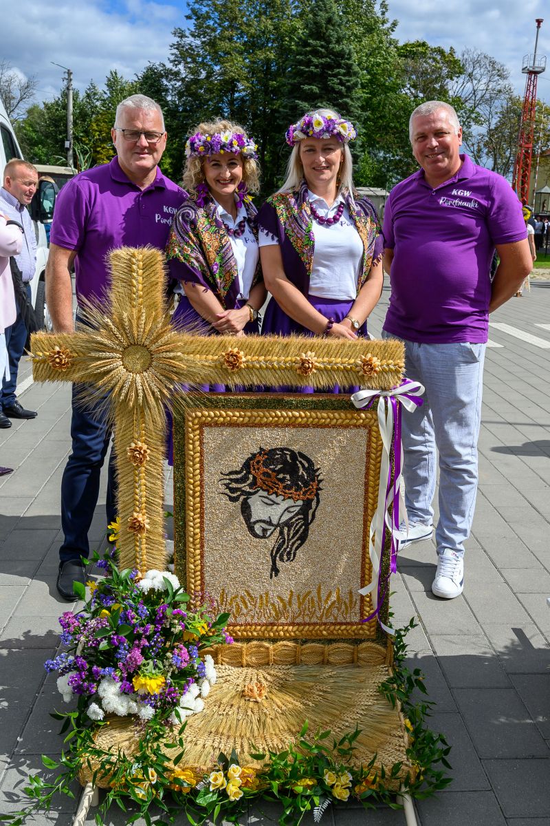 Grupa ludzi w fioletowych strojach stoi obok dzieła z kłosów i kwiatów. W tle drzewa i niebo. Centralny element kompozycji to religijny wizerunek z kłosów.