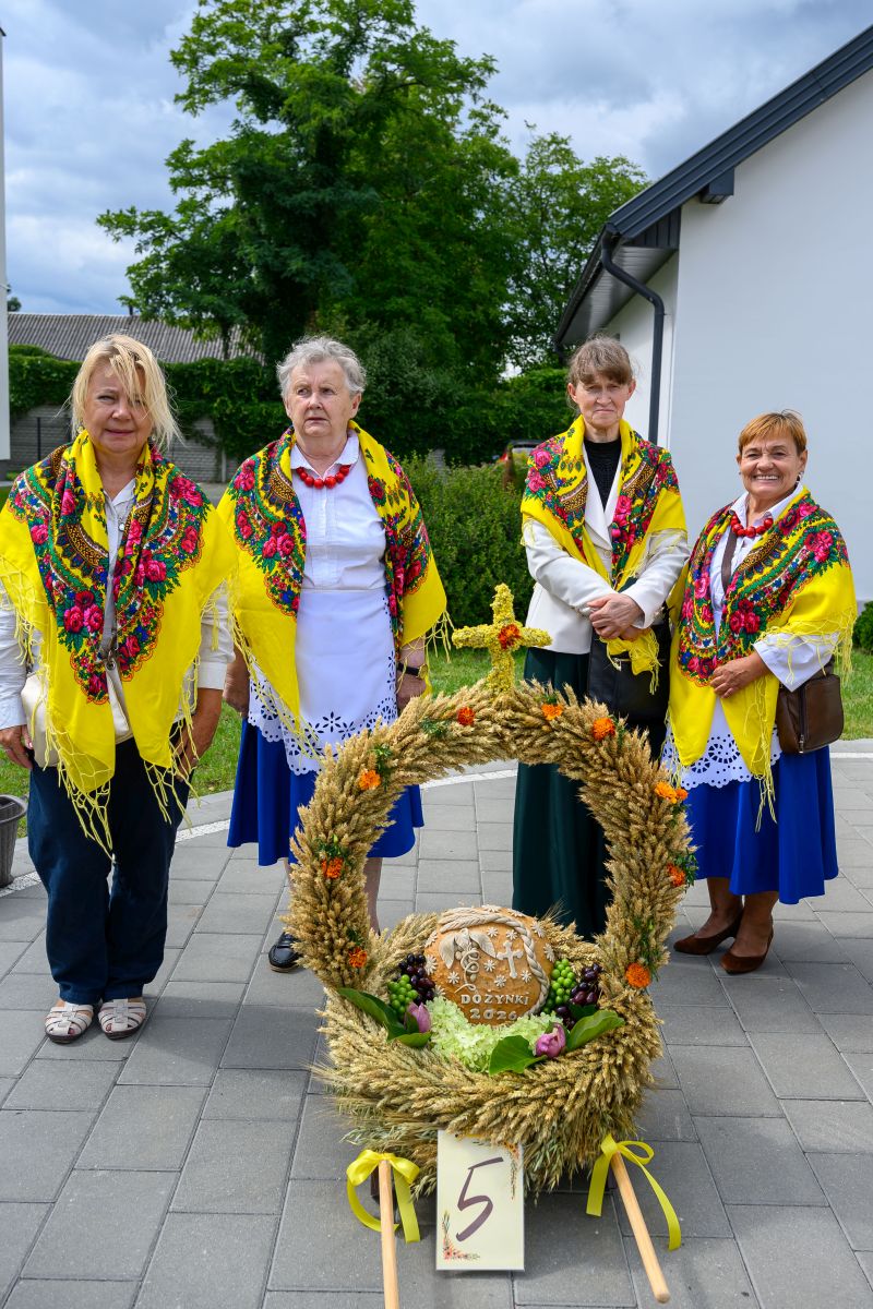 Cztery kobiety w ludowych strojach z kolorowymi chustami stoją za dożynkowym wieńcem z pszenicy i owoców. Tło to zielone krzewy i biała ściana budynku. Wieńce zdobią żółte i pomarańczowe akcenty.