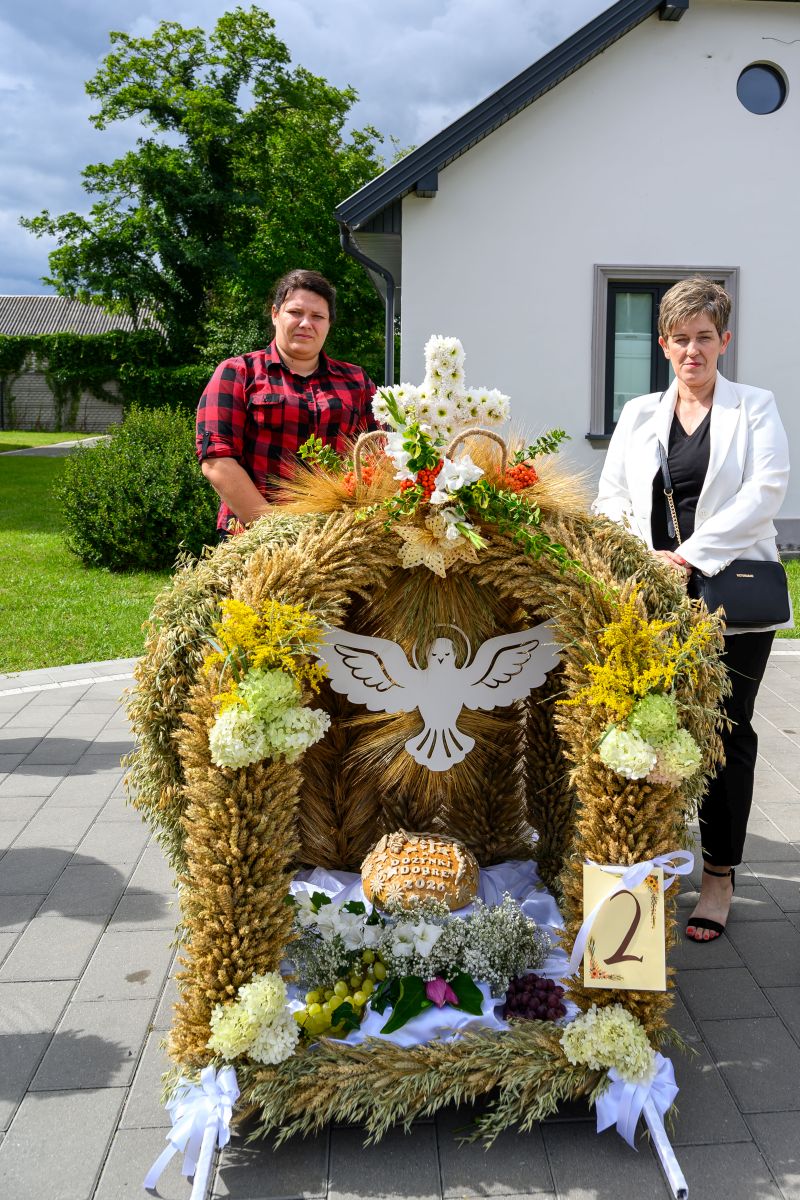 Dekoracja z kłosów zboża z białym ptakiem pośrodku, ozdobiona kwiatami i owocami. Przód domu w tle, zielone drzewa. Dwoje ludzi stoi obok, pogoda słoneczna.