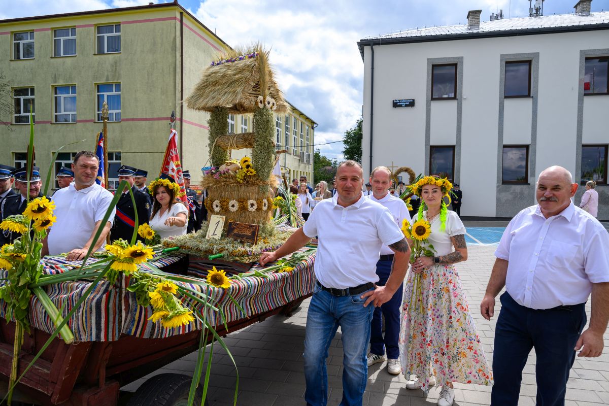 Grupa ludzi w białych koszulach przed wozem ozdobionym słomianą dekoracją i słonecznikami. W tle dwa budynki. Niebieskie niebo z chmurami.