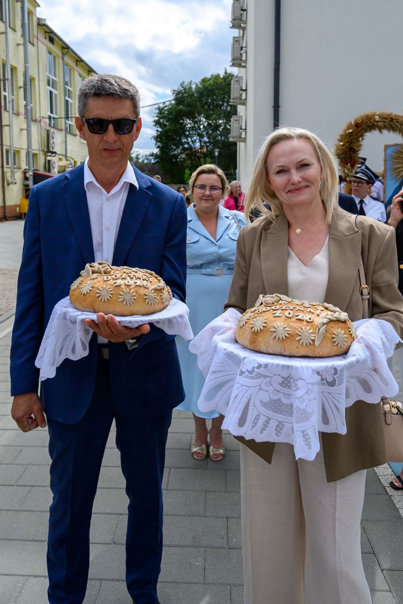 Dwie osoby trzymają dekoracyjne chleby na białych serwetach. Tło stanowią ludzie i budynek w jasnych kolorach. Atmosfera jest uroczysta.