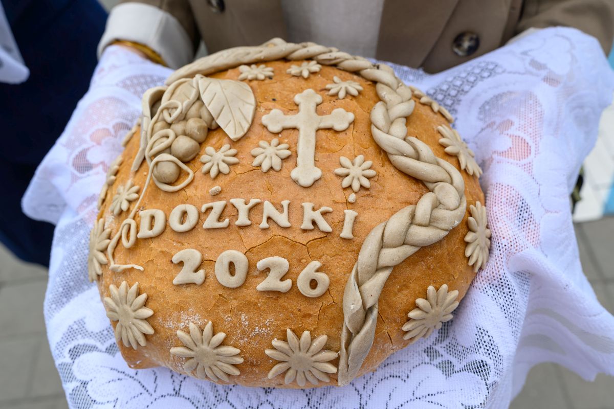 Na zdjęciu widać bochen chleba ozdobiony napisem DOŻYNKI 2026, kwiatami i krzyżem. Tło jest białe, koronkowe. Bochen trzymany jest w rękach na obrusem.