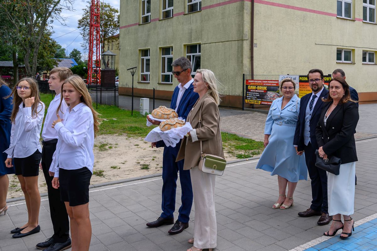 Grupa ludzi stoi przed budynkiem, niektórzy w białych koszulach, z chlebem na tacy. Tło jest zielone z drzewami. Budynek w kolorze kremowym z oknami.