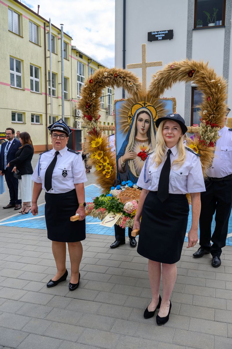 Na zdjęciu dwie kobiety w białych koszulach i ciemnych spódnicach w procesji z wizerunkiem Maryi. Tło stanowi budynek szkoły.