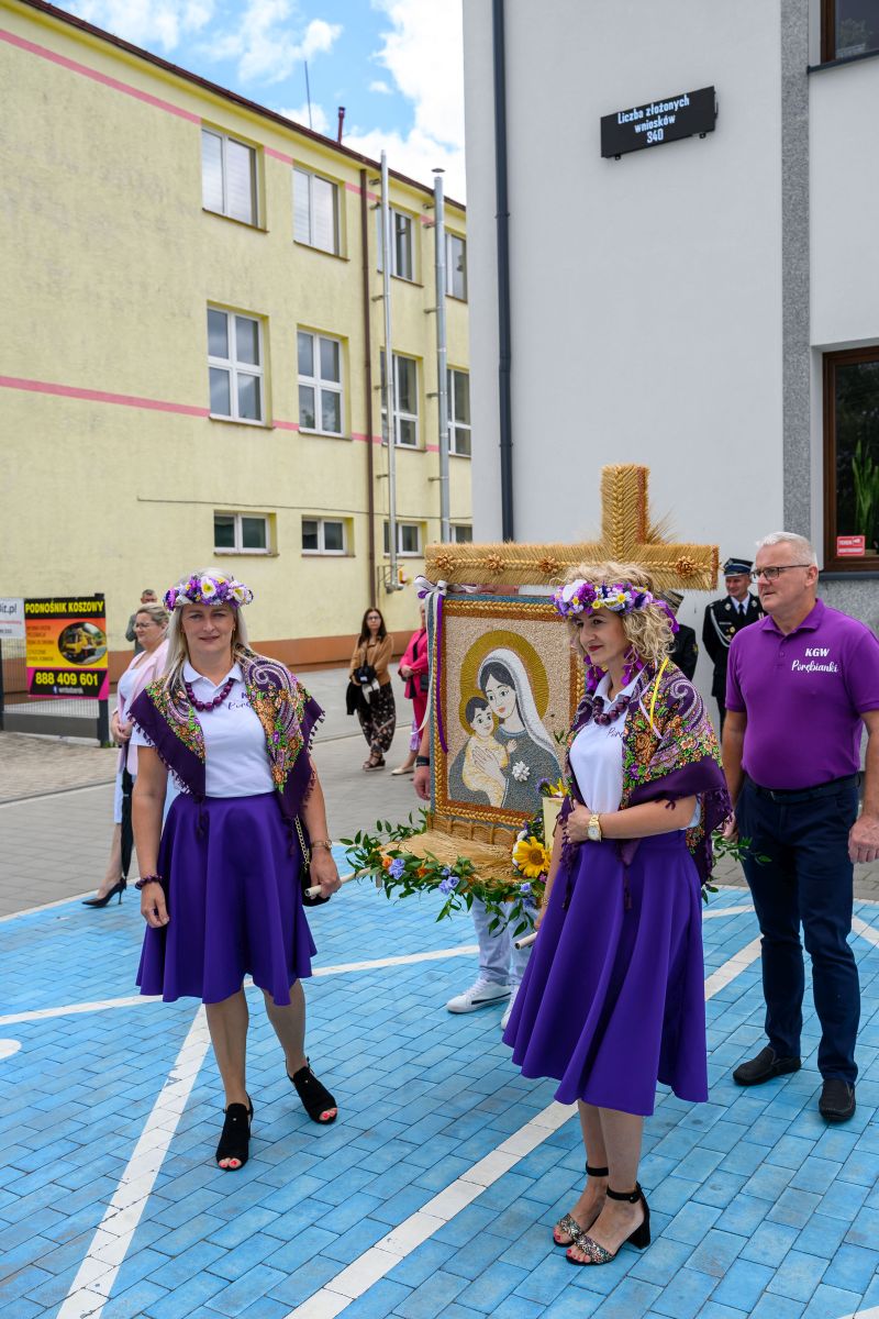 Święto religijne z osobami w fioletowych strojach i wieńcem. Tło to budynek w żółci i szarości. Na chodniku niebieskie płytki.