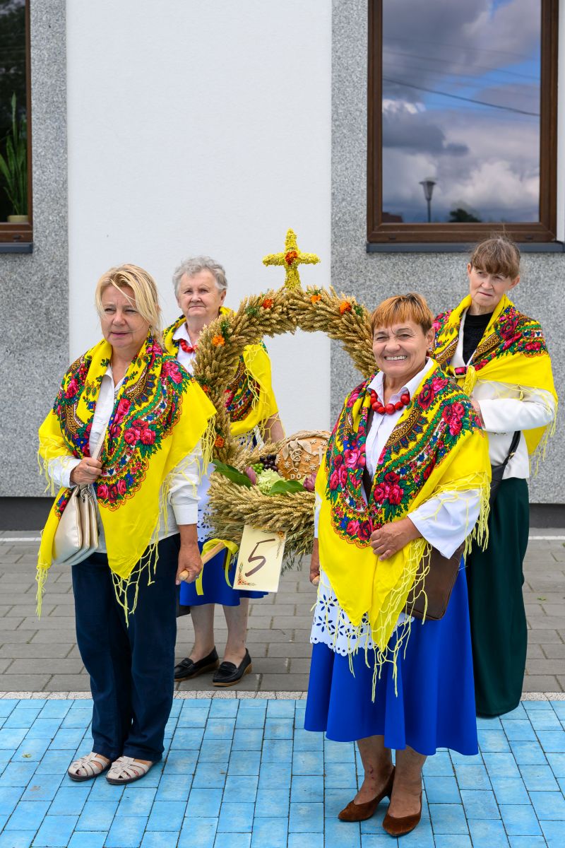 Na zdjęciu cztery kobiety w kolorowych chustach trzymają dożynkowy wieniec zbożowy. Tło to biało-szary budynek. Kobiety ubrane są w biel i odcienie niebieskiego.