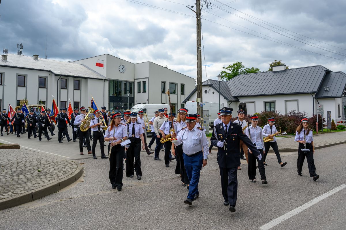 Parada strażaków w mundurach maszerujących obok budynku z flagą Polski. W tle pochmurne niebo. Widoczne instrumenty muzyczne.